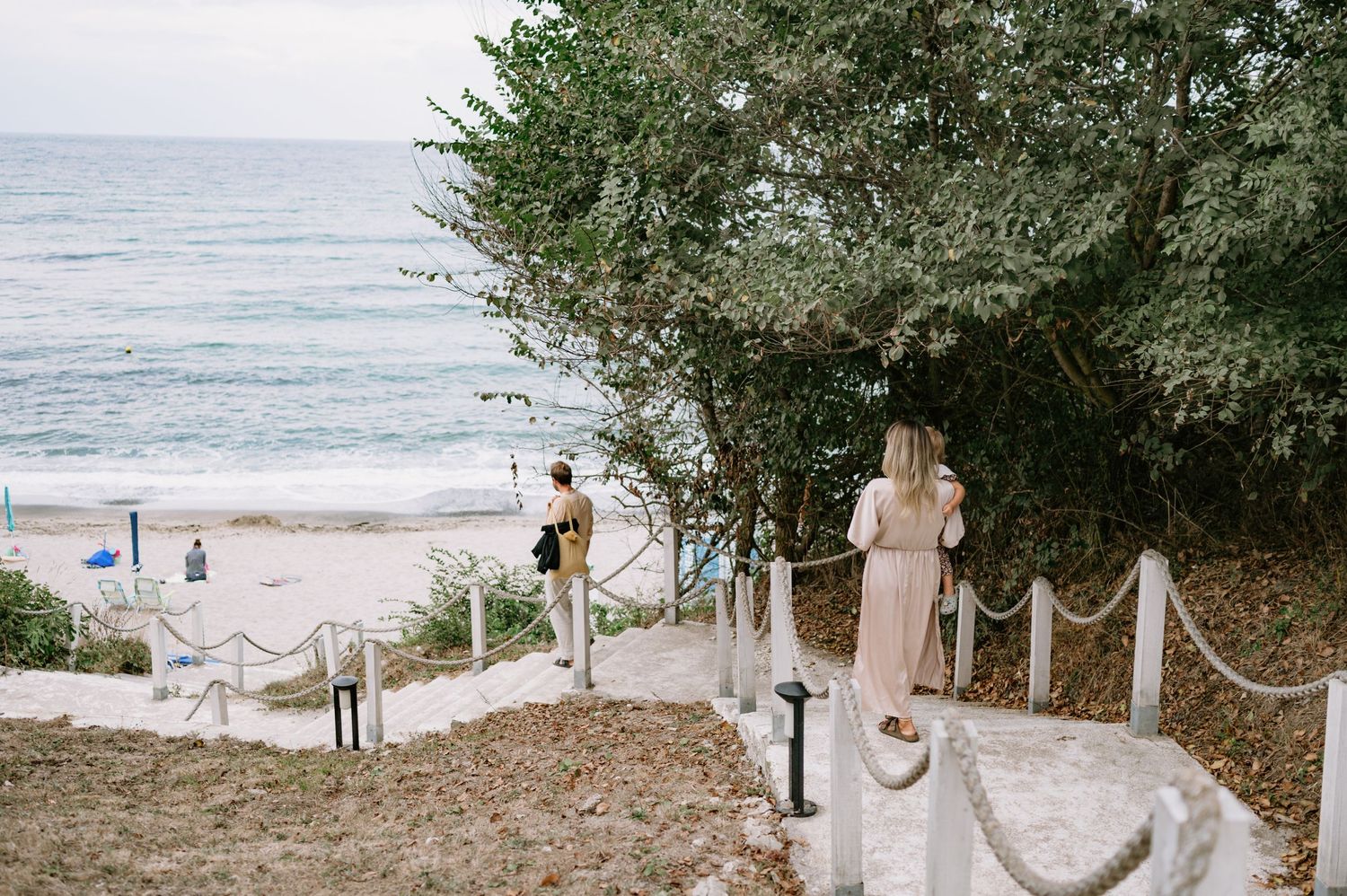 Couple walking down coastal stairs toward a sandy beach, framed by lush green trees and ocean waves.