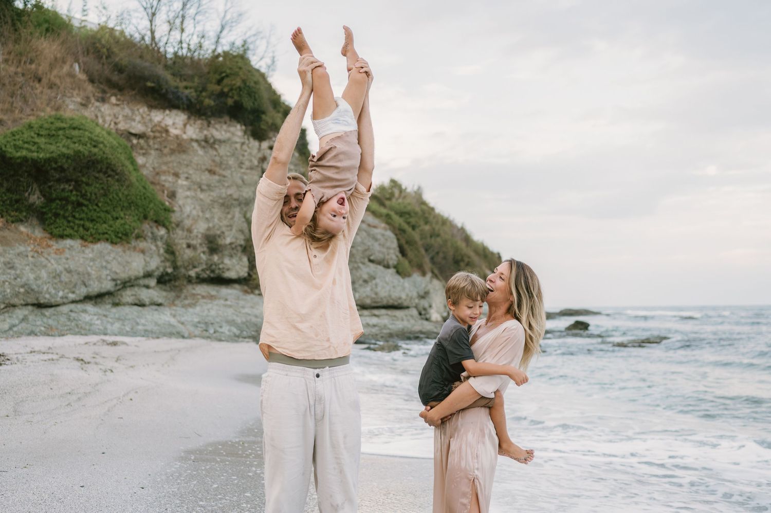 Family enjoying a beach day, father lifting baby overhead while mother holds toddler by rocky cliffs.