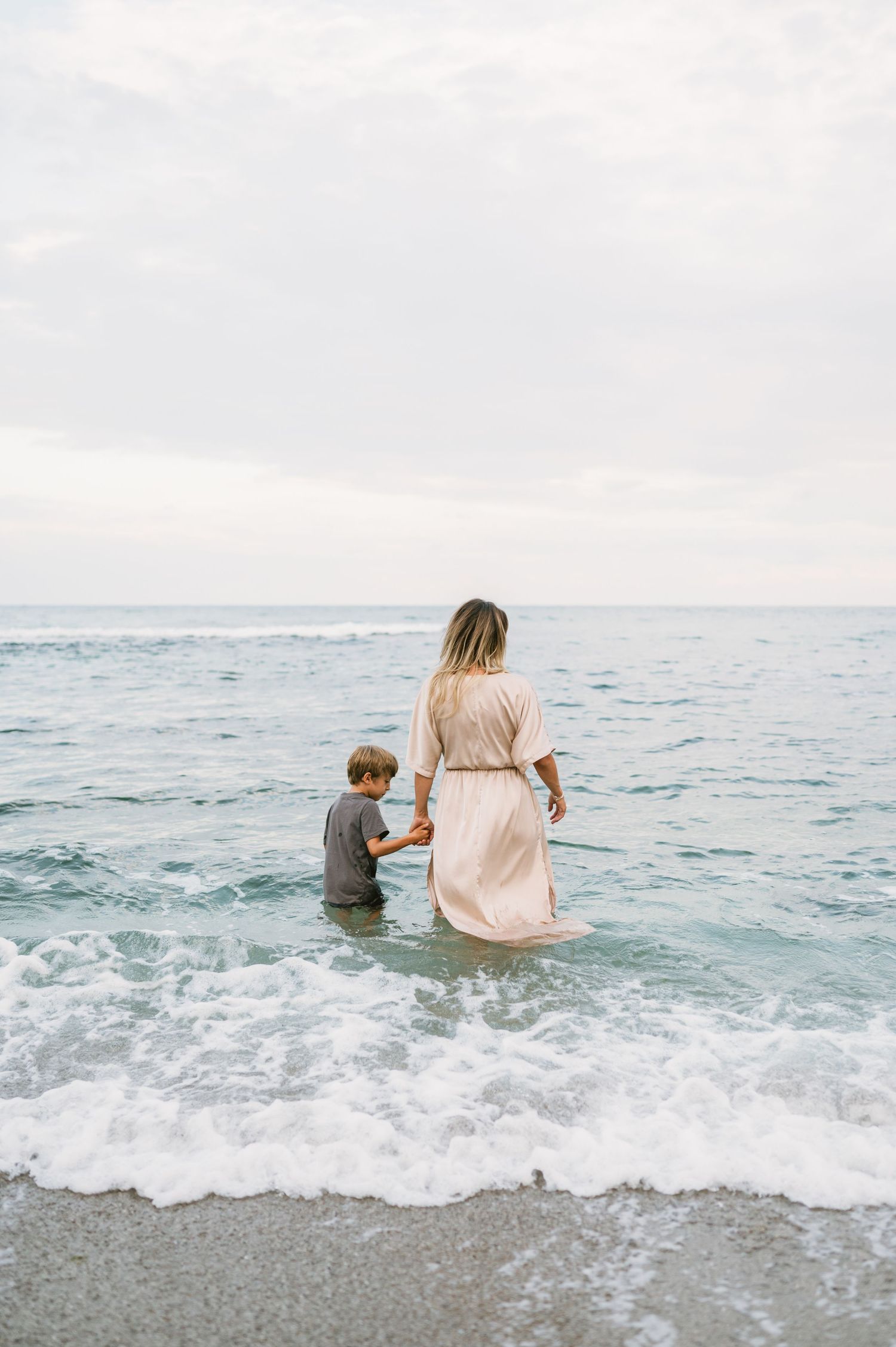 A woman in a pink dress holds hands with a young boy wading in ocean waves on a cloudy beach day.