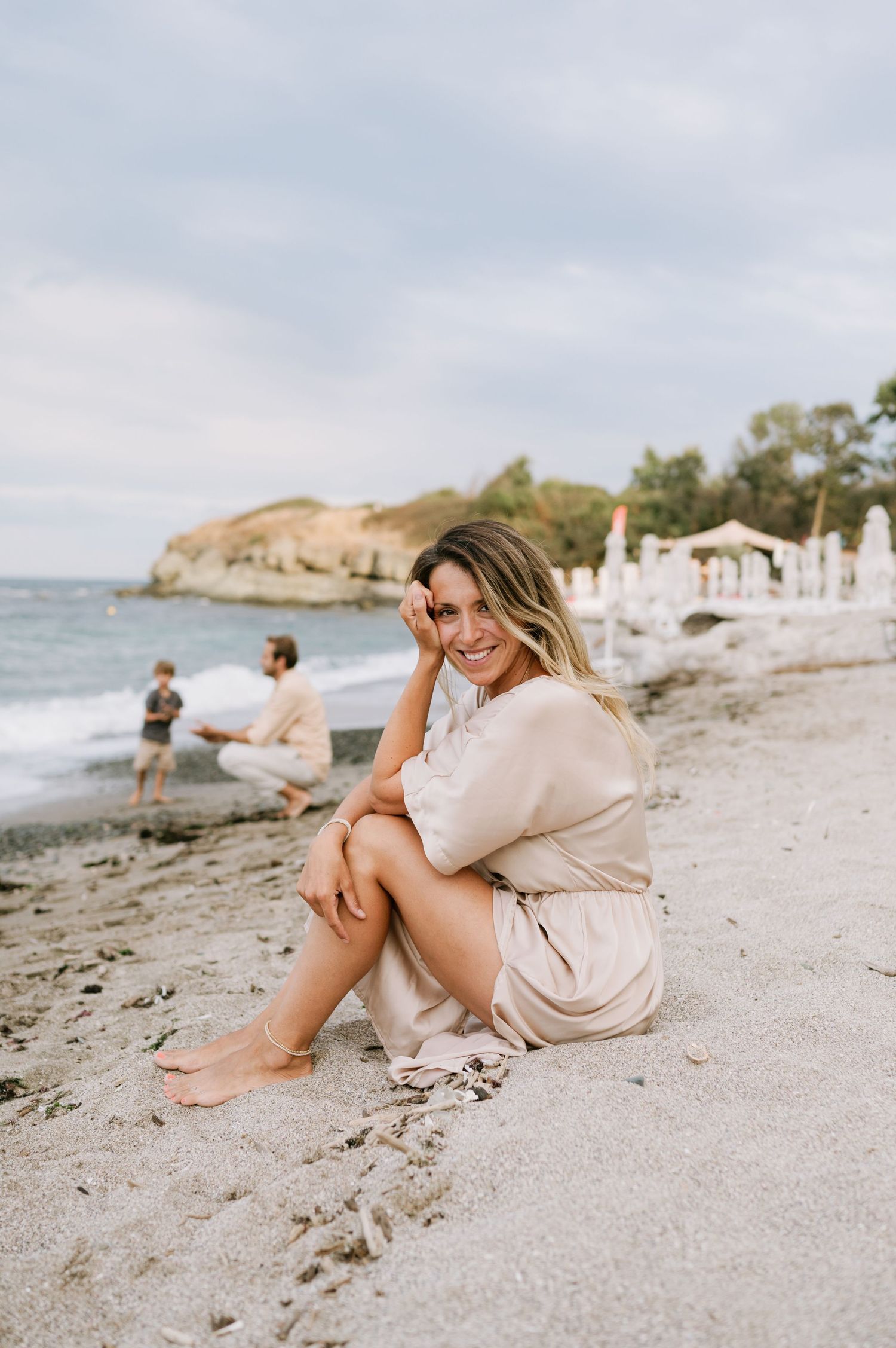Woman in beige dress sitting barefoot on sandy beach, smiling at camera with ocean and umbrellas in background.