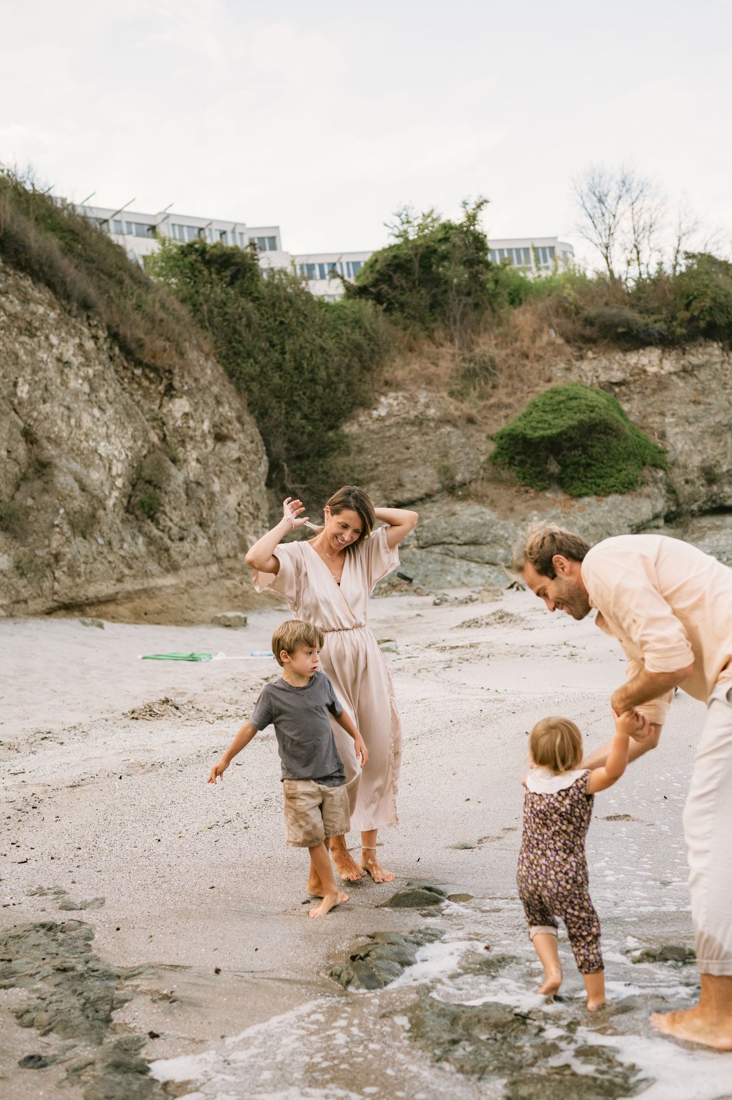 Family of four playing barefoot on a sandy beach near rocky cliffs, with waves washing ashore on a cloudy day.