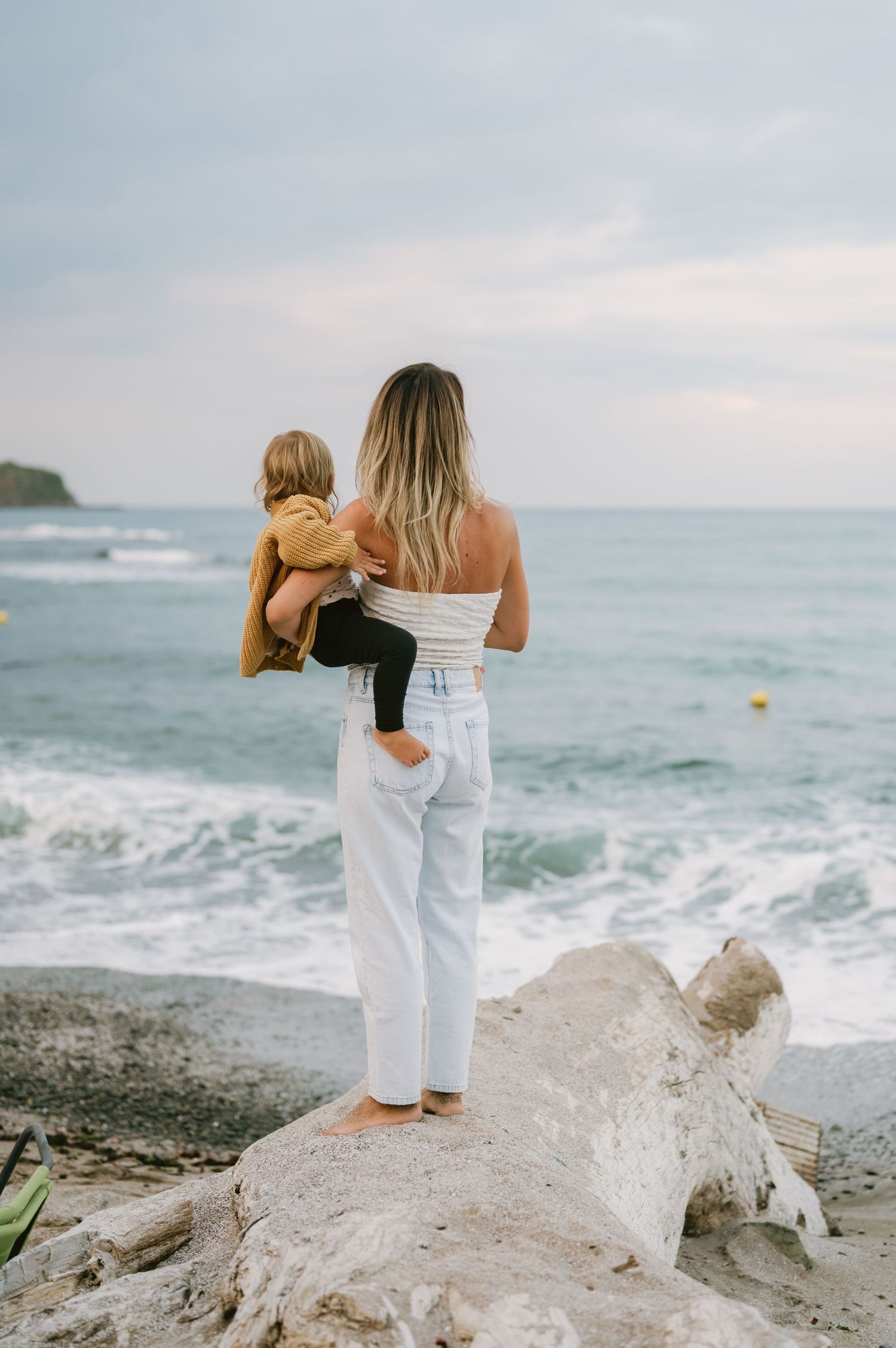 A woman holding a toddler stands on driftwood at a beach, both facing the ocean waves under a cloudy sky.