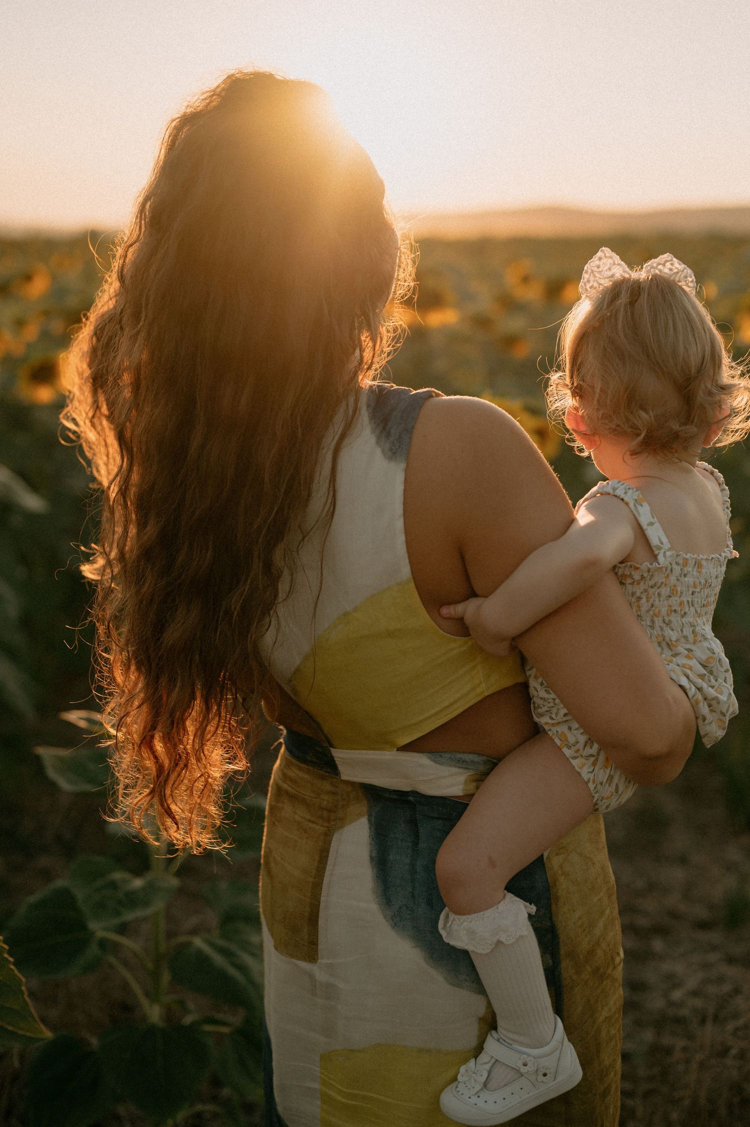 Mother holding toddler in sunflower field at golden hour sunset, viewed from behind with long flowing hair.