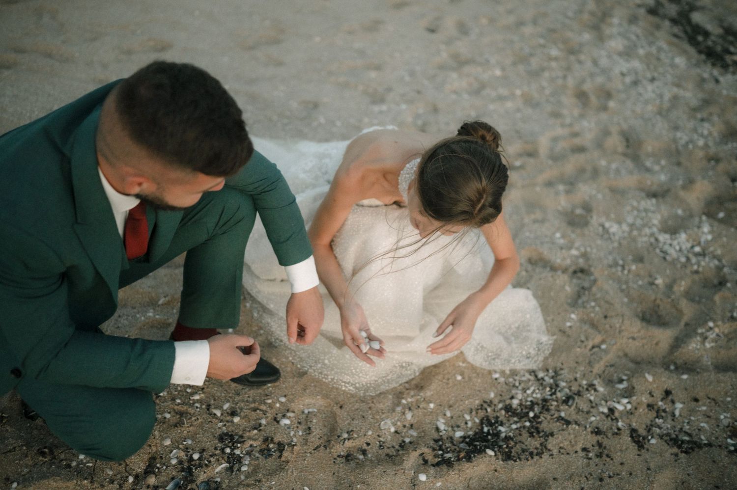 Bride in white gown and groom in green suit kneel on sandy beach, examining shells together.
