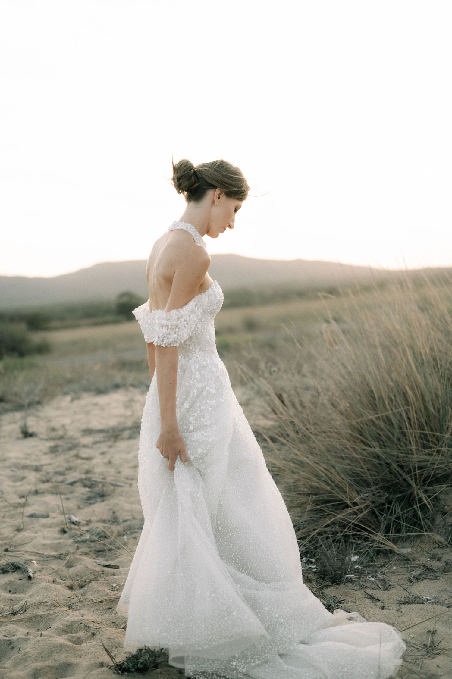 Bride in off-shoulder white lace gown standing on sandy dunes at sunset with golden grass backdrop.