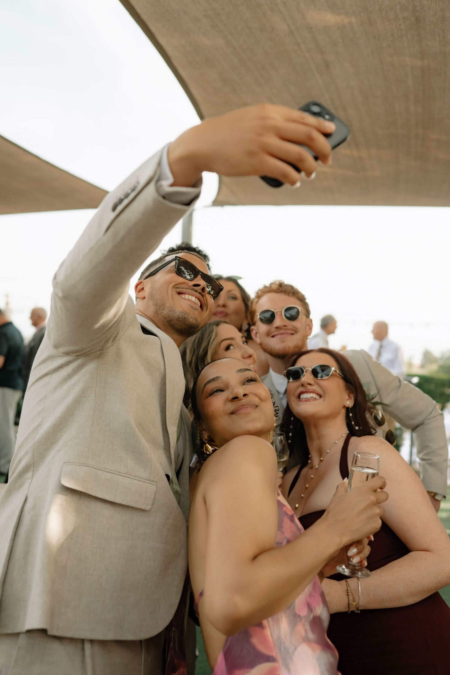 Group of friends in sunglasses taking a selfie at an outdoor wedding reception on a sunny day.