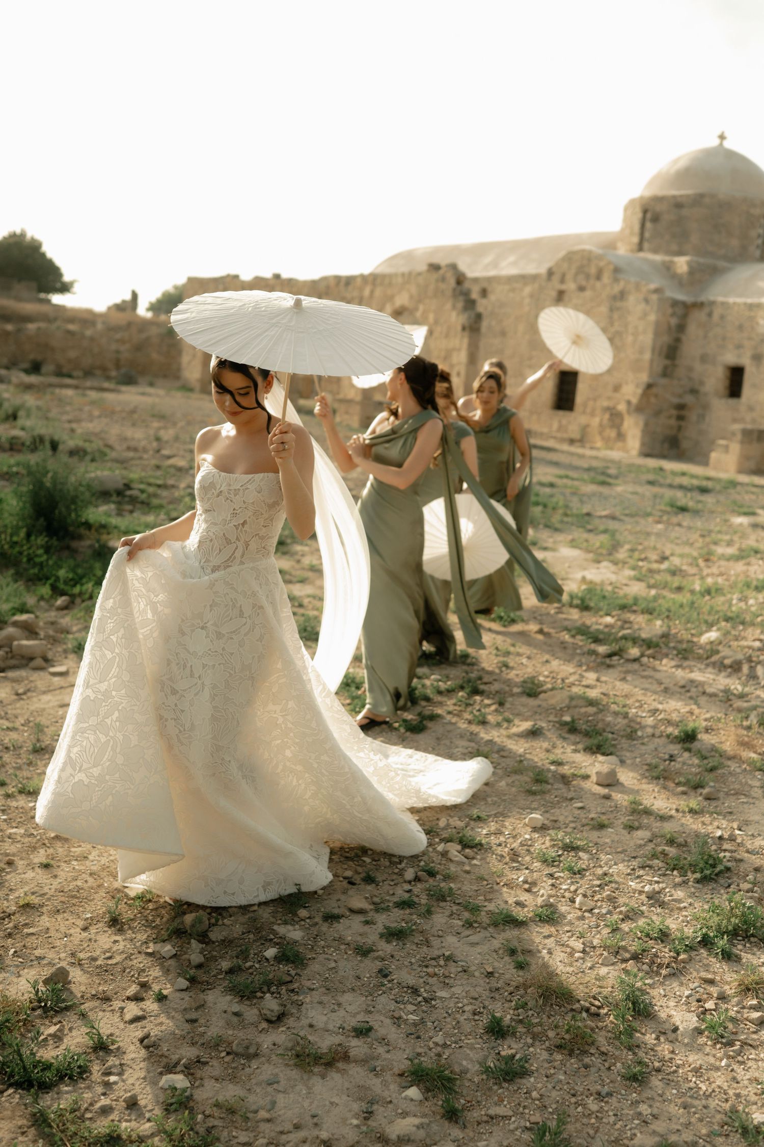 Bride in lace gown with parasol leads bridesmaids in sage dresses past ancient stone ruins on sunny day.