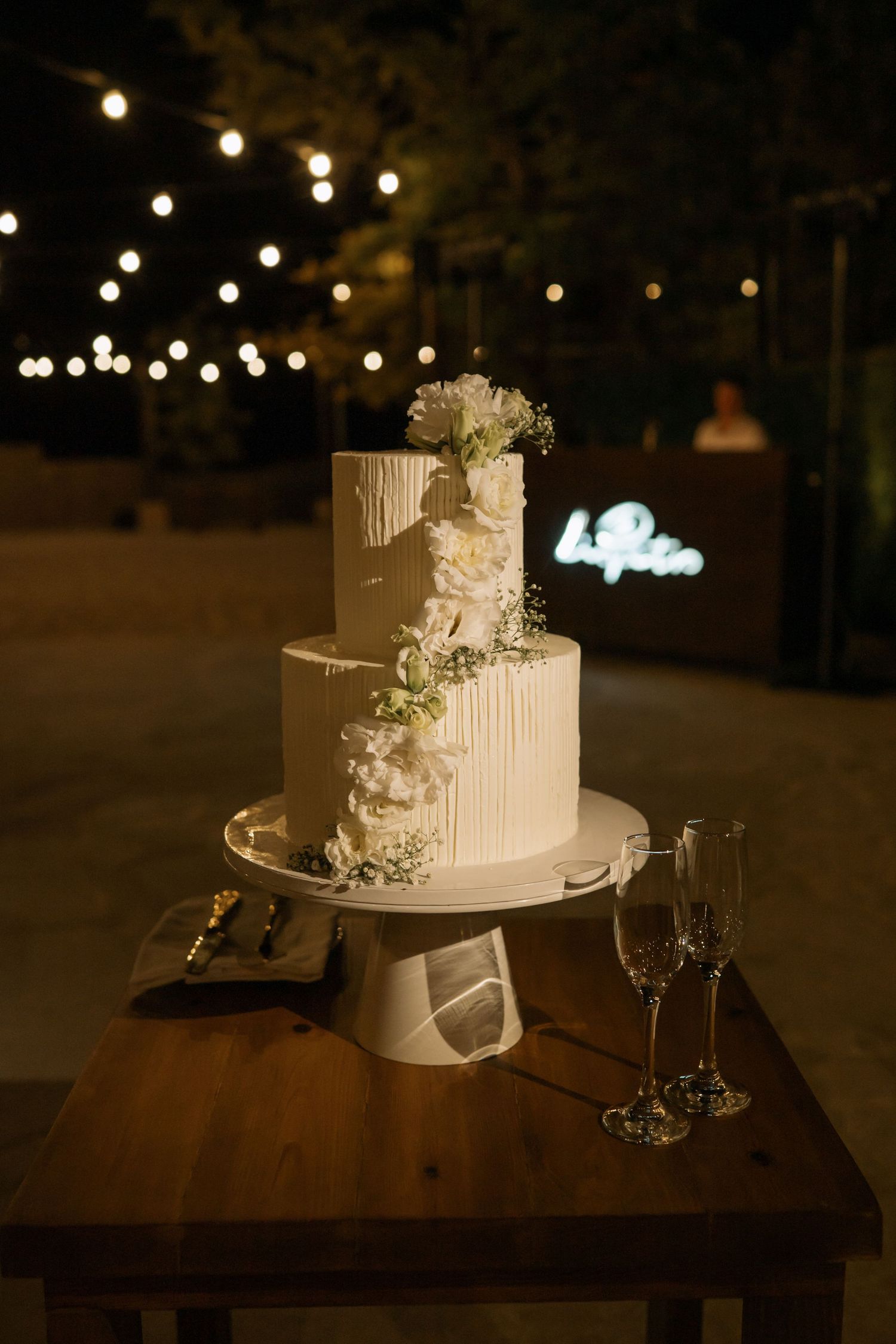 Elegant two-tier white wedding cake with floral decor on wooden table, champagne flutes, string lights at night.