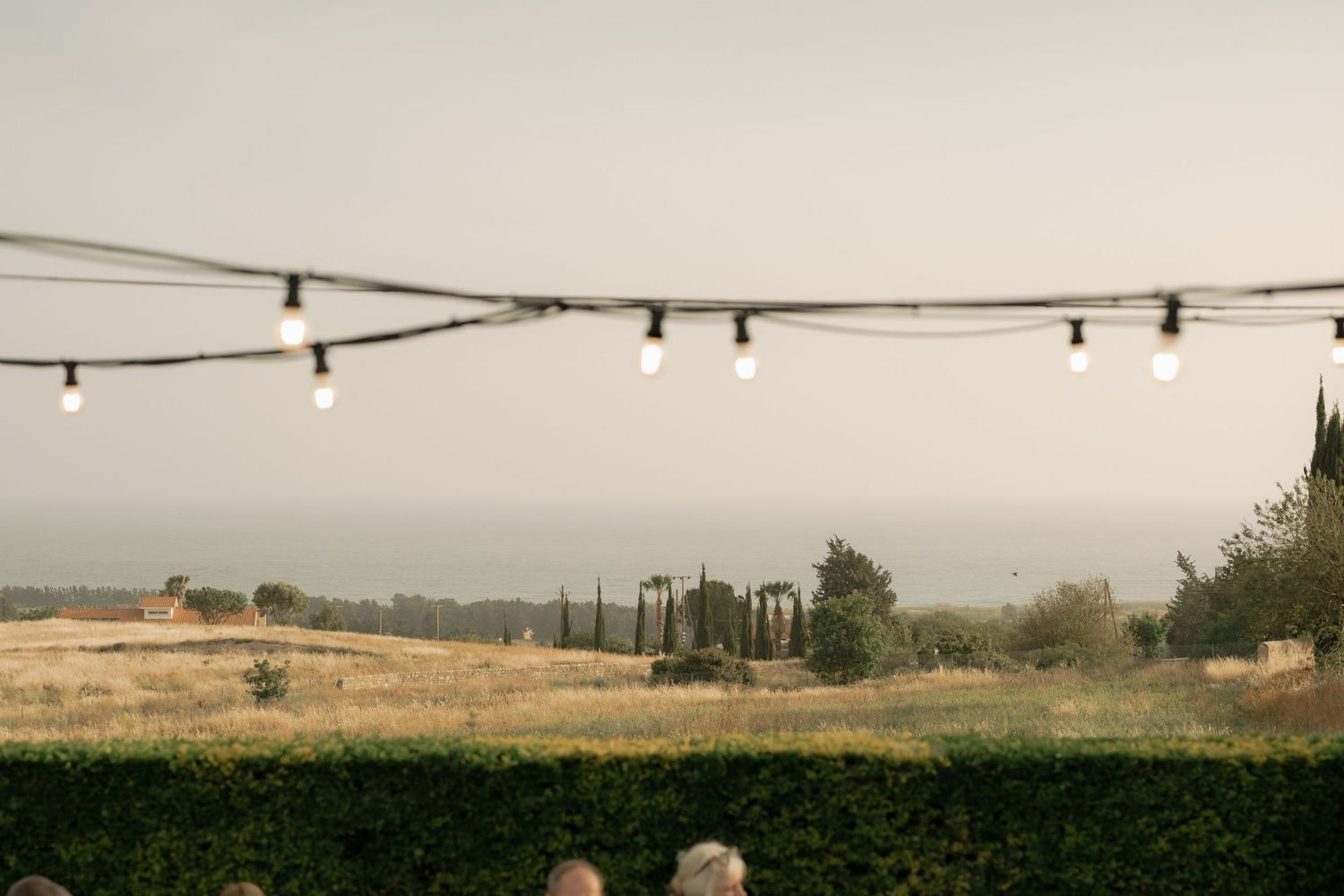 String lights hang over a rustic Tuscan countryside landscape with cypress trees and golden fields at dusk.