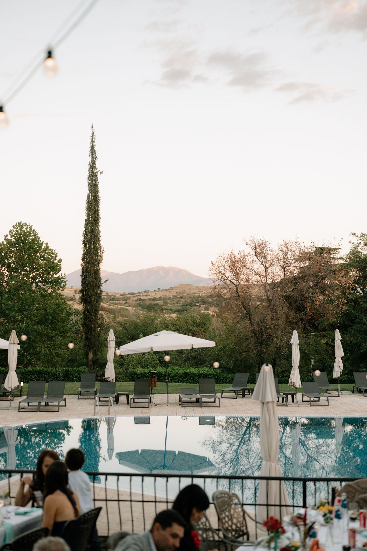 People enjoying a poolside gathering at dusk with tall cypress trees and mountains in the background.