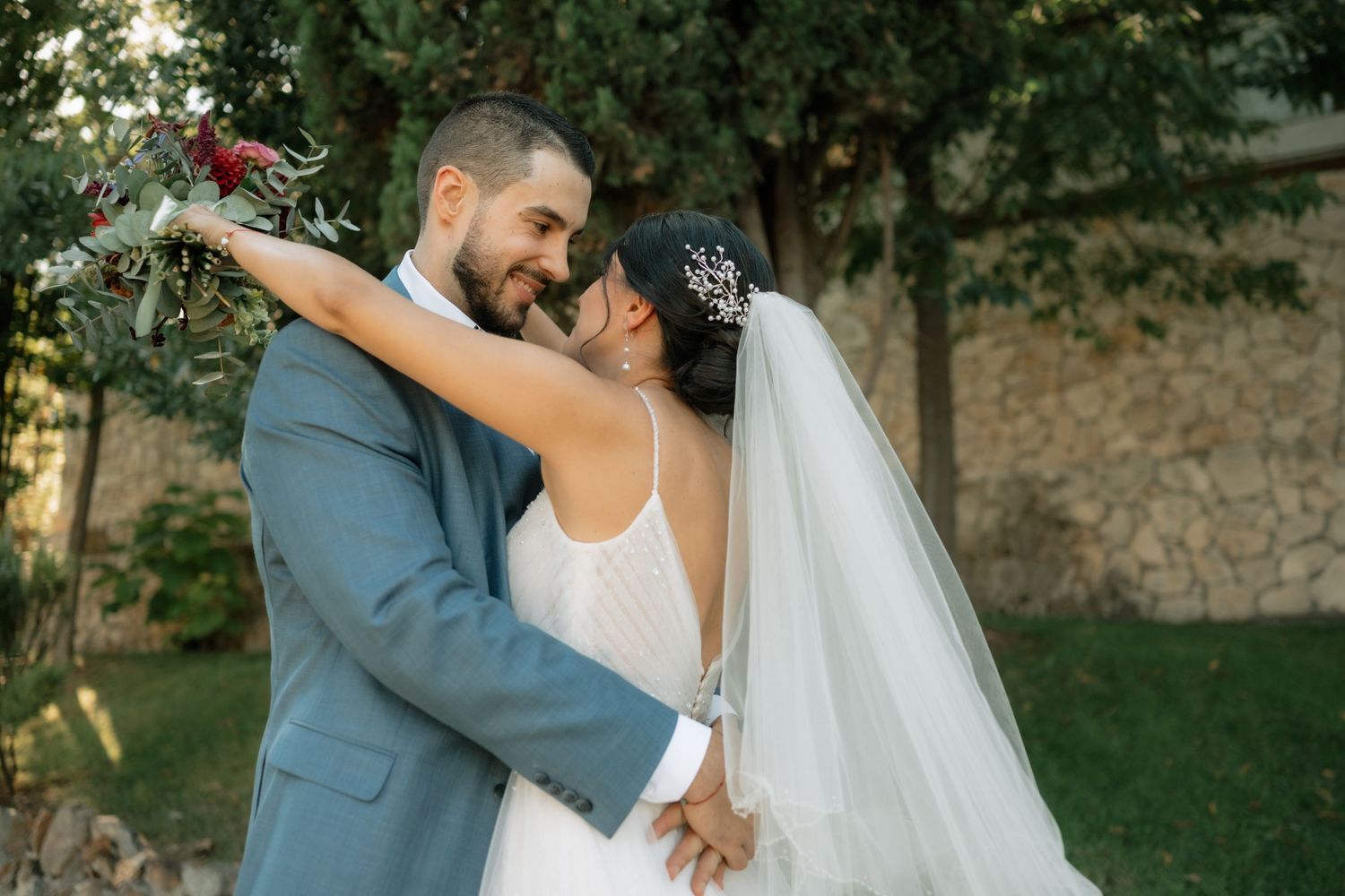 A romantic newlywed couple embraces outdoors in a garden setting with the bride wearing a flowing veil and white dress.