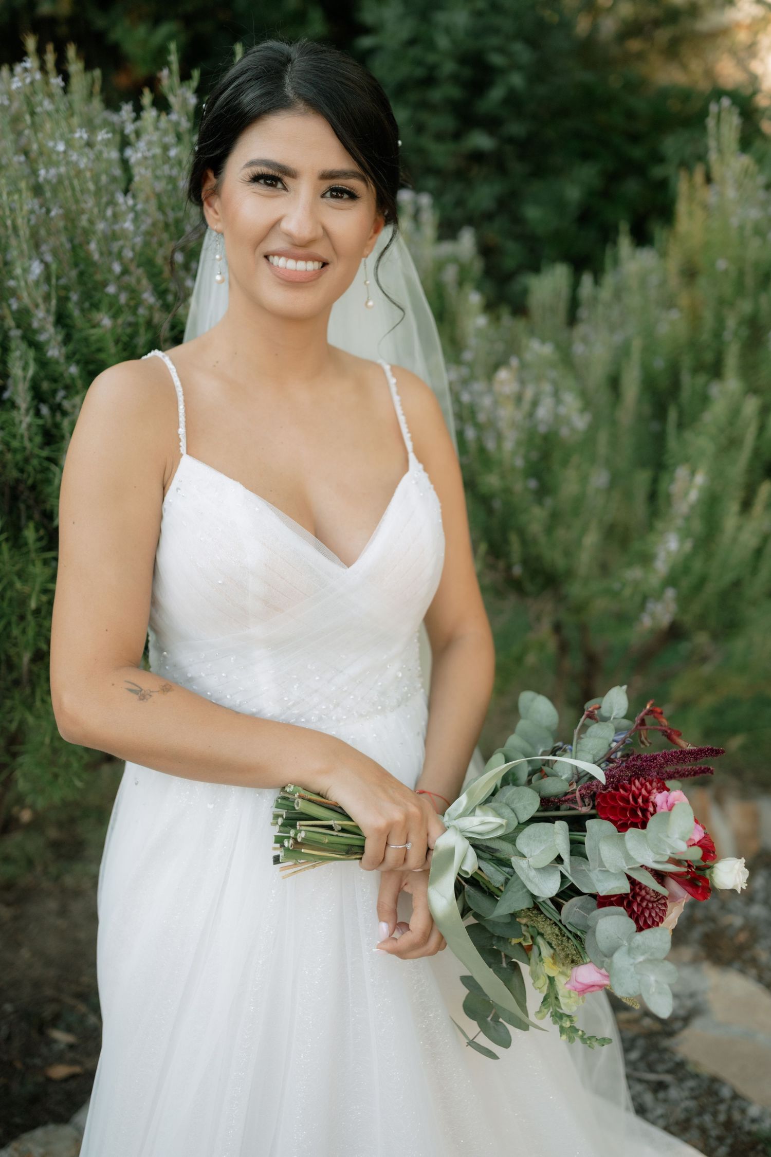 A bride in a white wedding dress poses with her eucalyptus and burgundy floral bouquet in an outdoor garden setting.