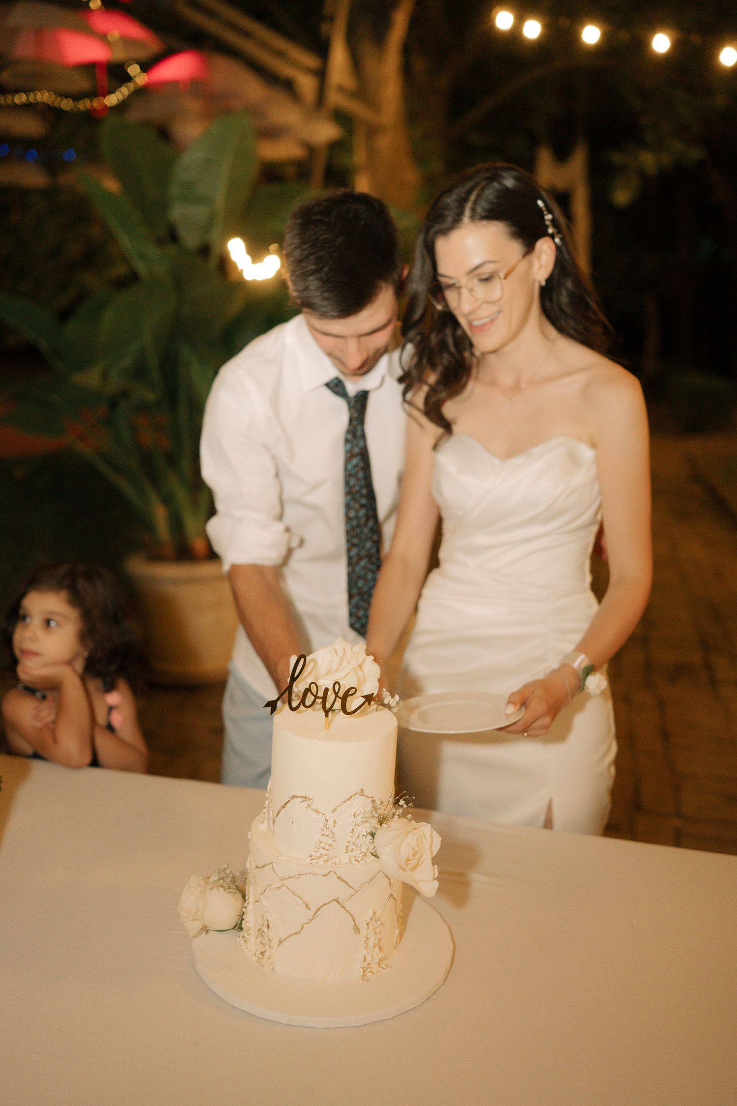 A couple in wedding attire cuts into an elegant white wedding cake under warm lighting at an evening reception.