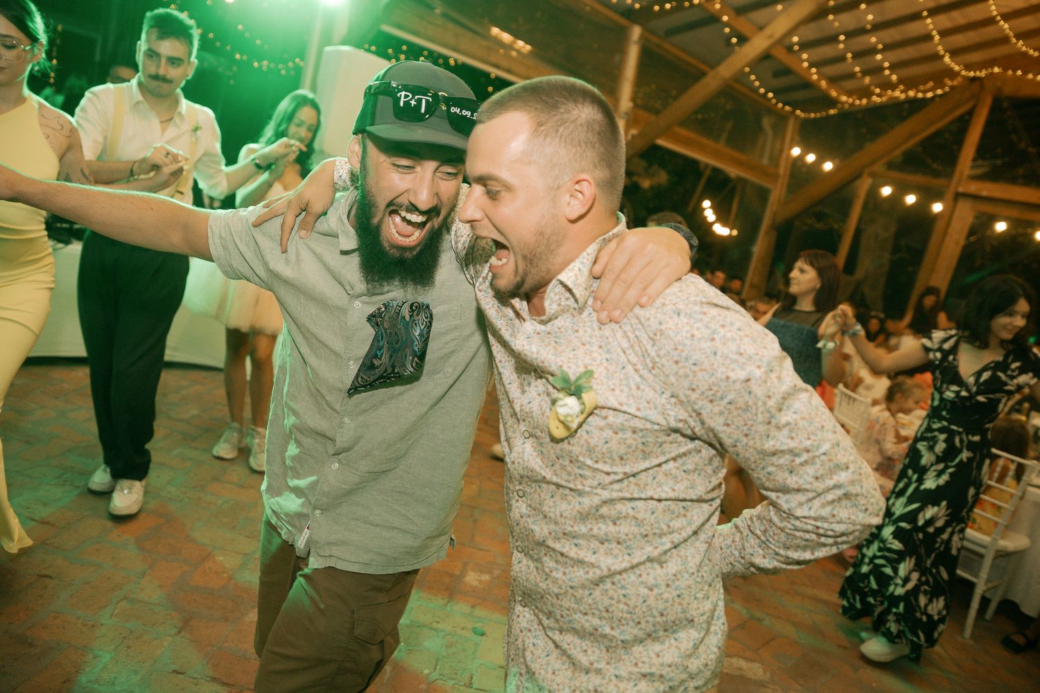 Two friends dancing and laughing together at a reception venue with string lights and guests in the background.