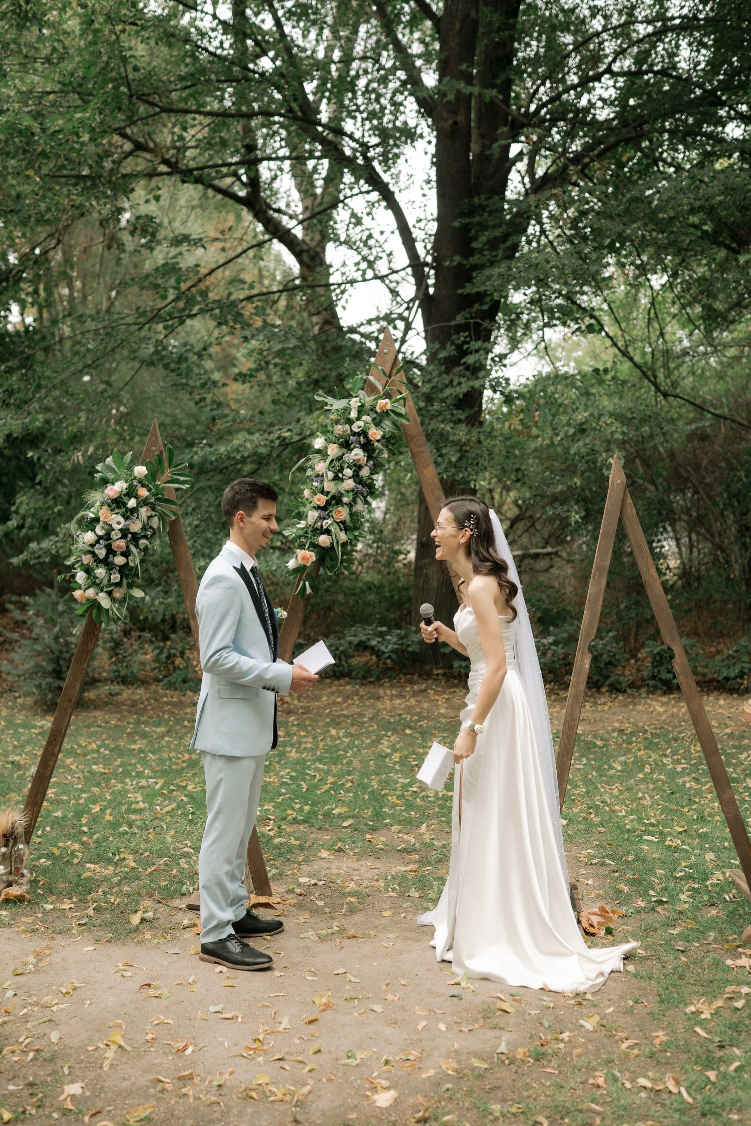 A couple in wedding attire shares a playful moment on wooden swings in a wooded outdoor setting with fallen leaves.