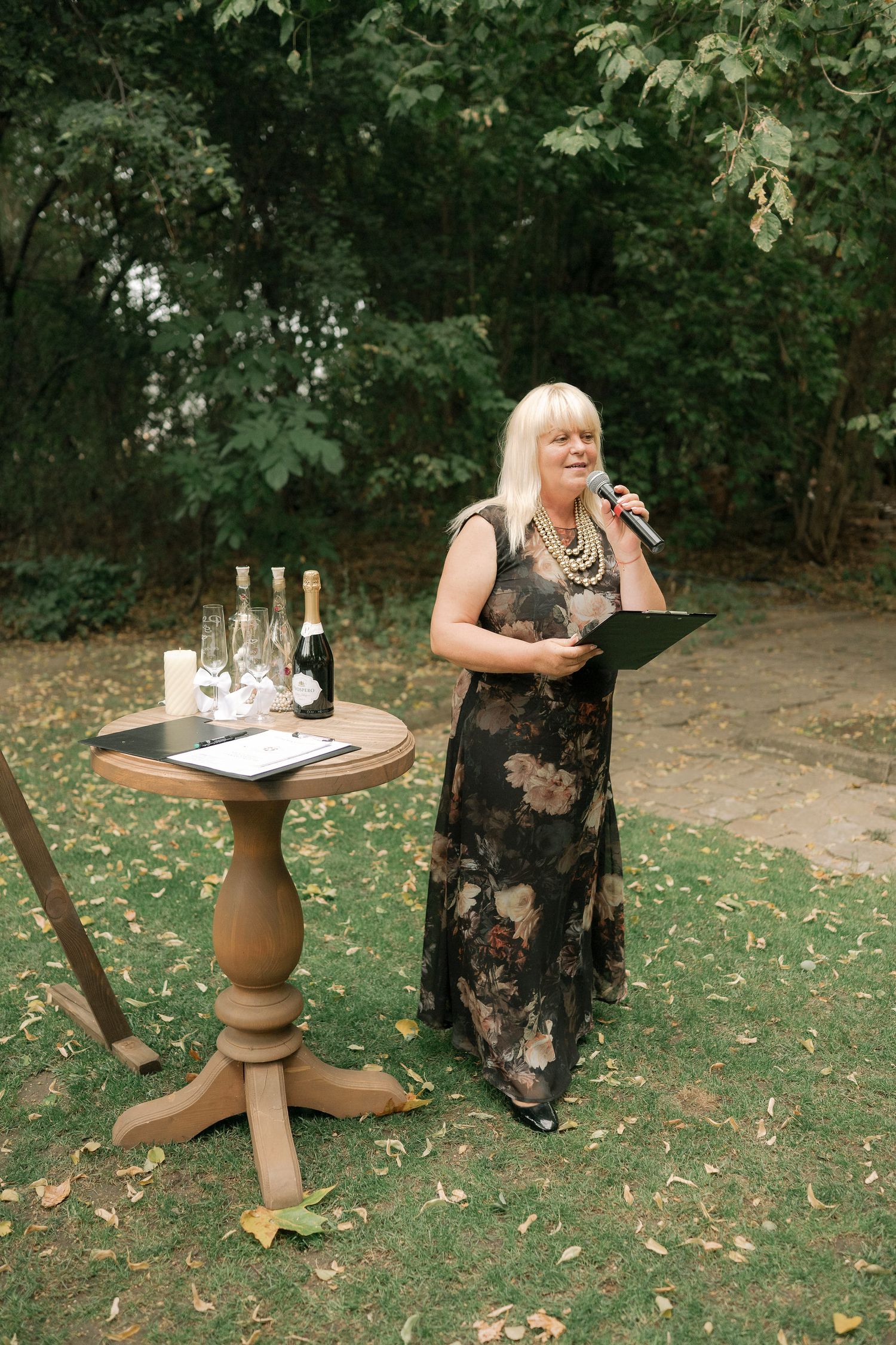 A person in a floral dress stands near a decorative pedestal table in a garden setting.