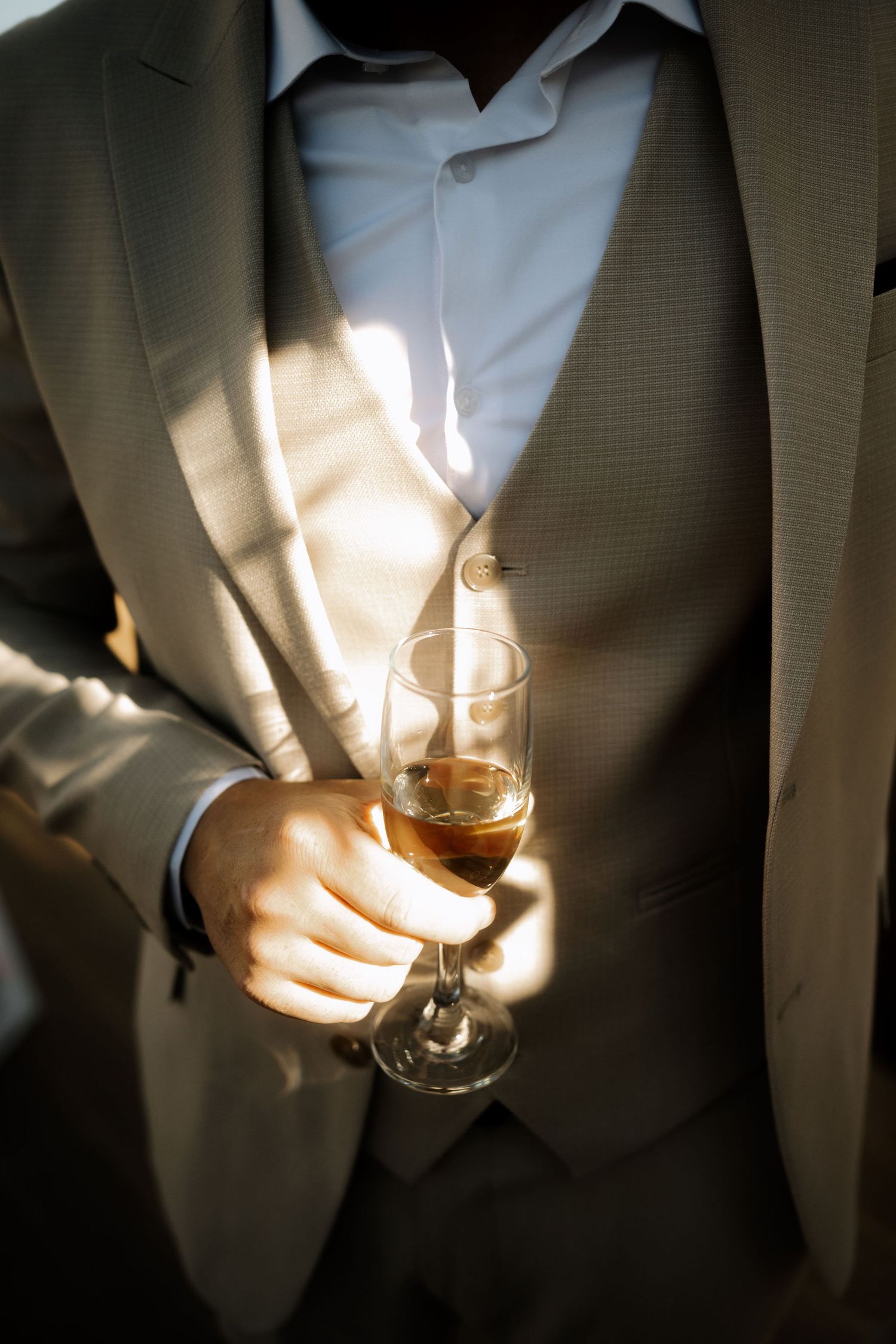 A person in a light gray suit holds a glass of wine with sunlight casting dramatic shadows across their torso.