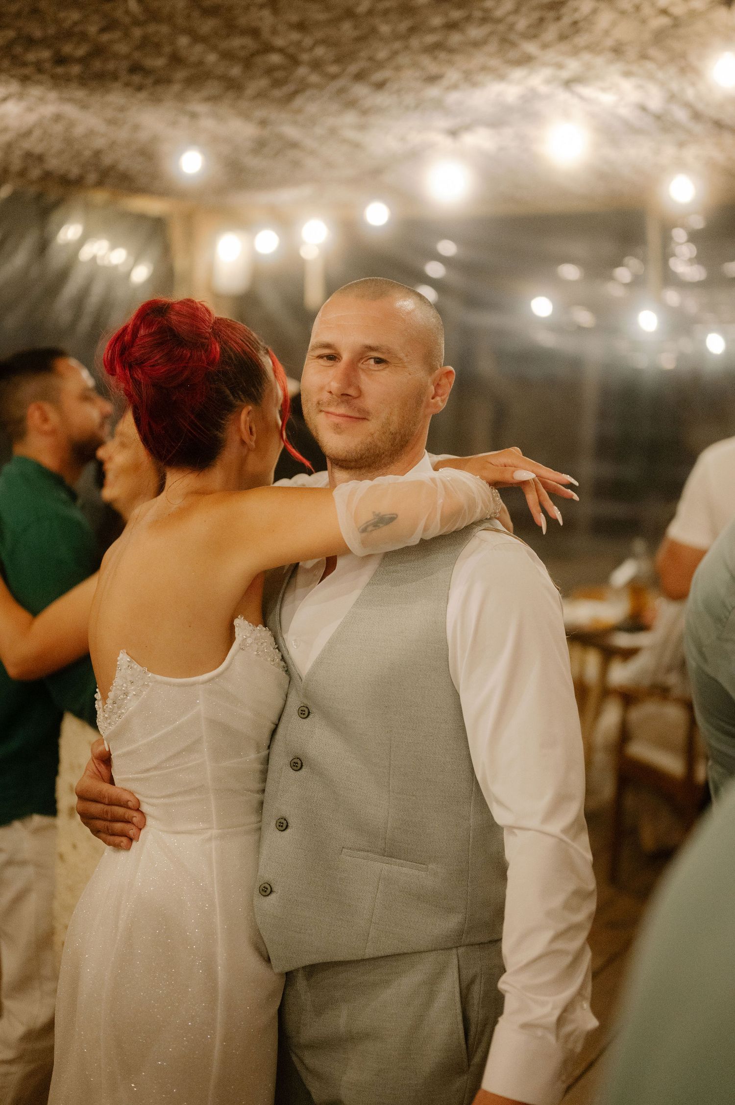 A couple shares a romantic dance moment under warm string lights at an evening wedding reception.