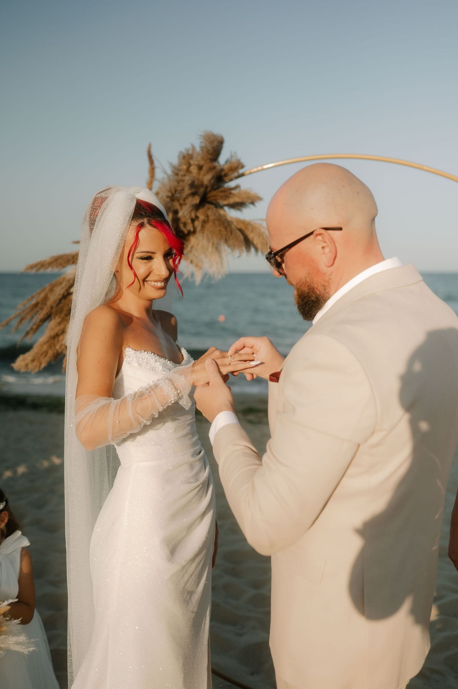A couple exchanges rings during a beach wedding ceremony with pampas grass decor and ocean backdrop at sunset.