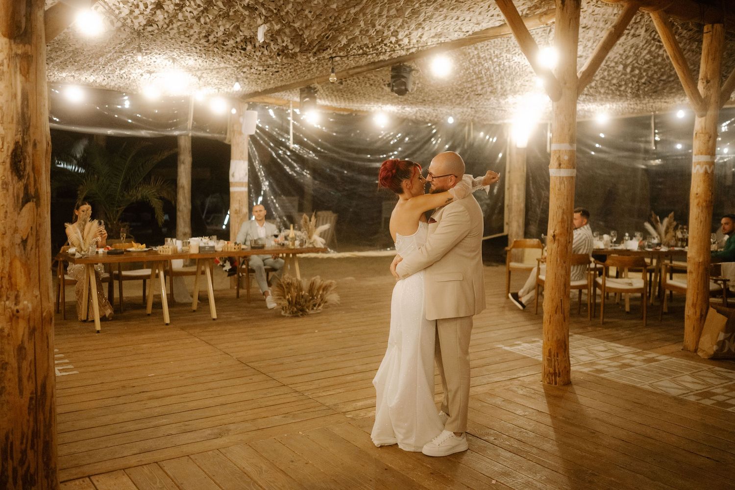 A string of warm lights illuminate a rustic wooden deck dance floor at an outdoor evening reception.