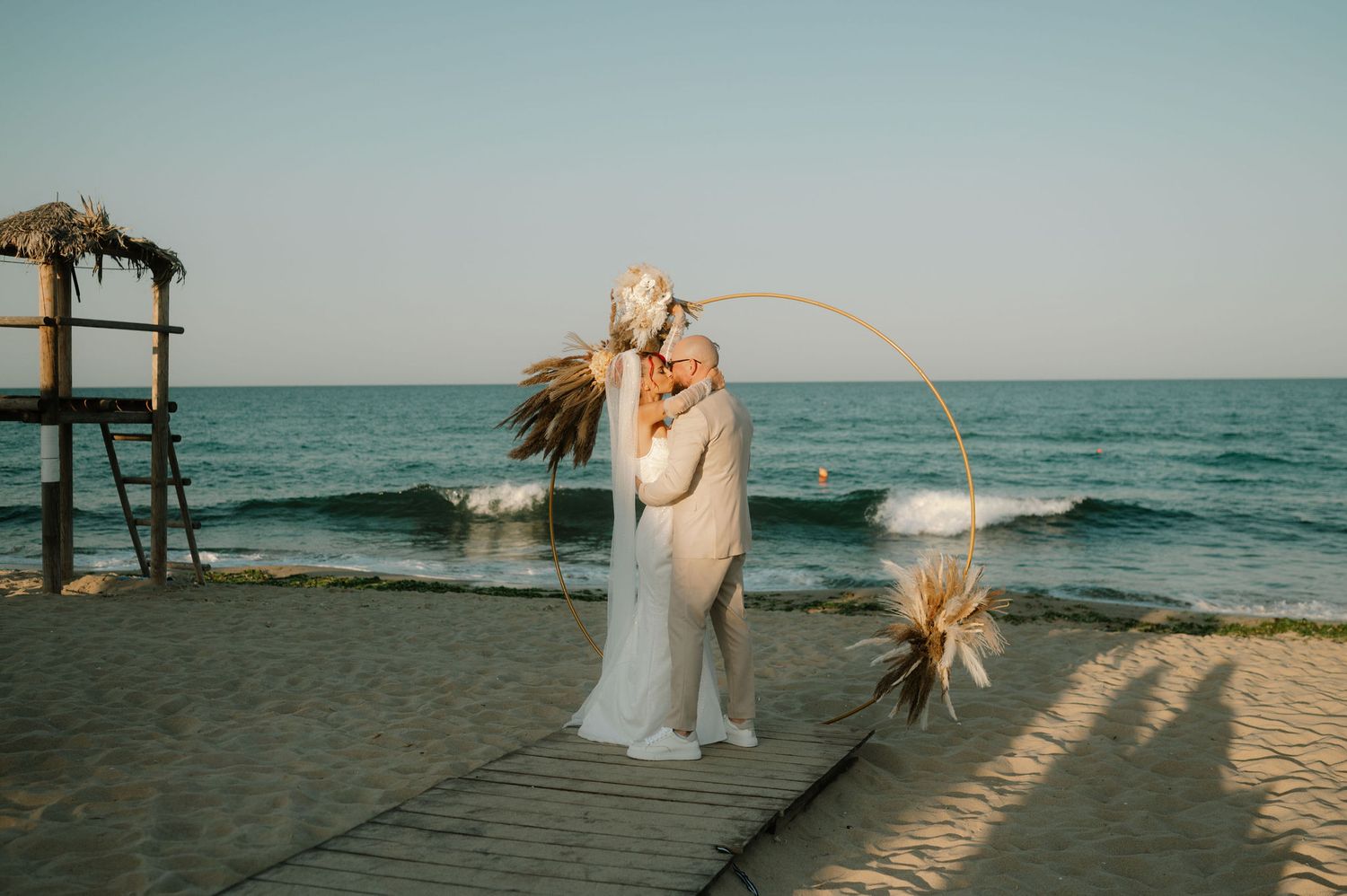 A romantic beach wedding ceremony setup at sunset with a wooden arch and tropical floral decorations by the ocean.