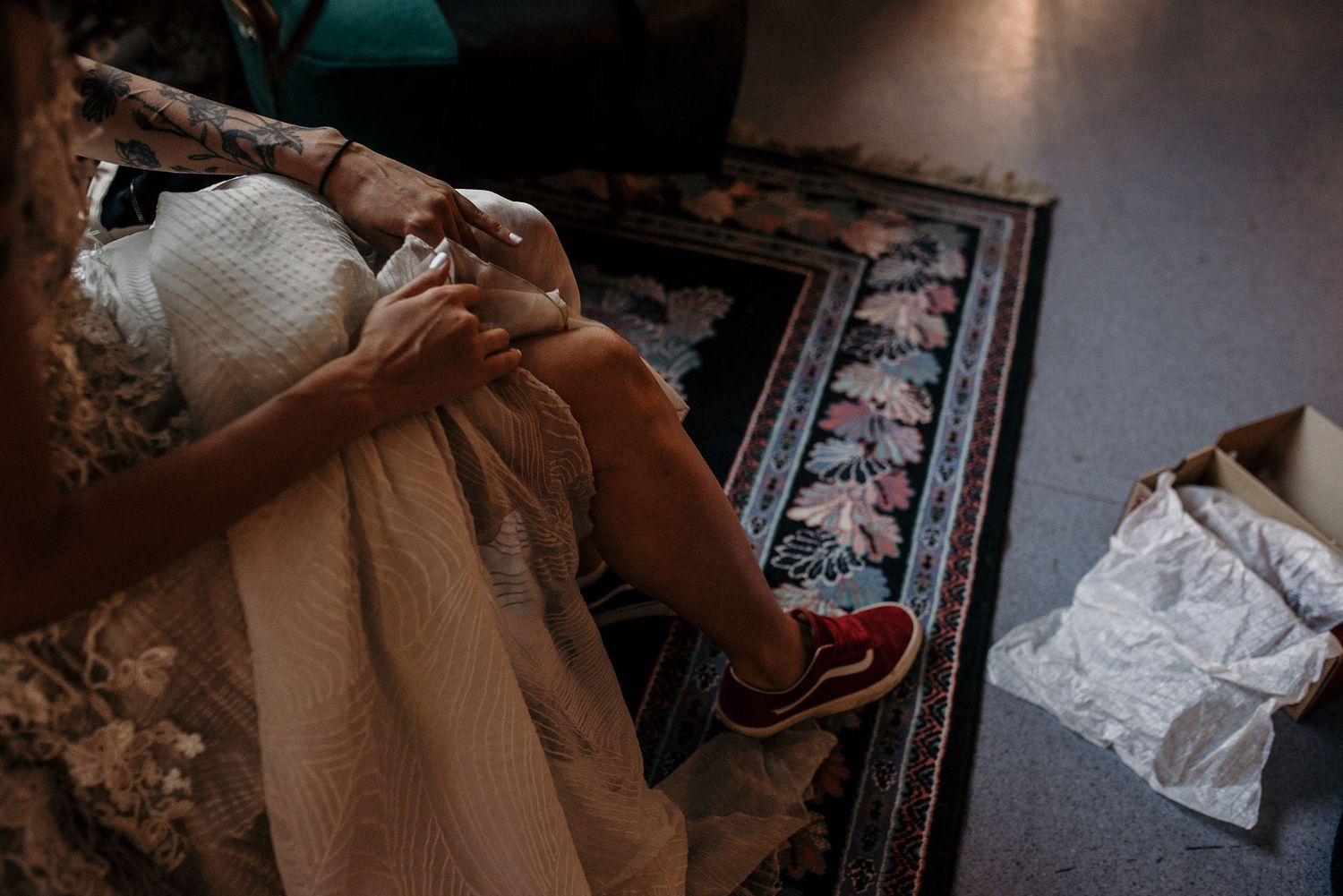 Hands holding knitting needles work on a cream-colored project against a patterned rug.