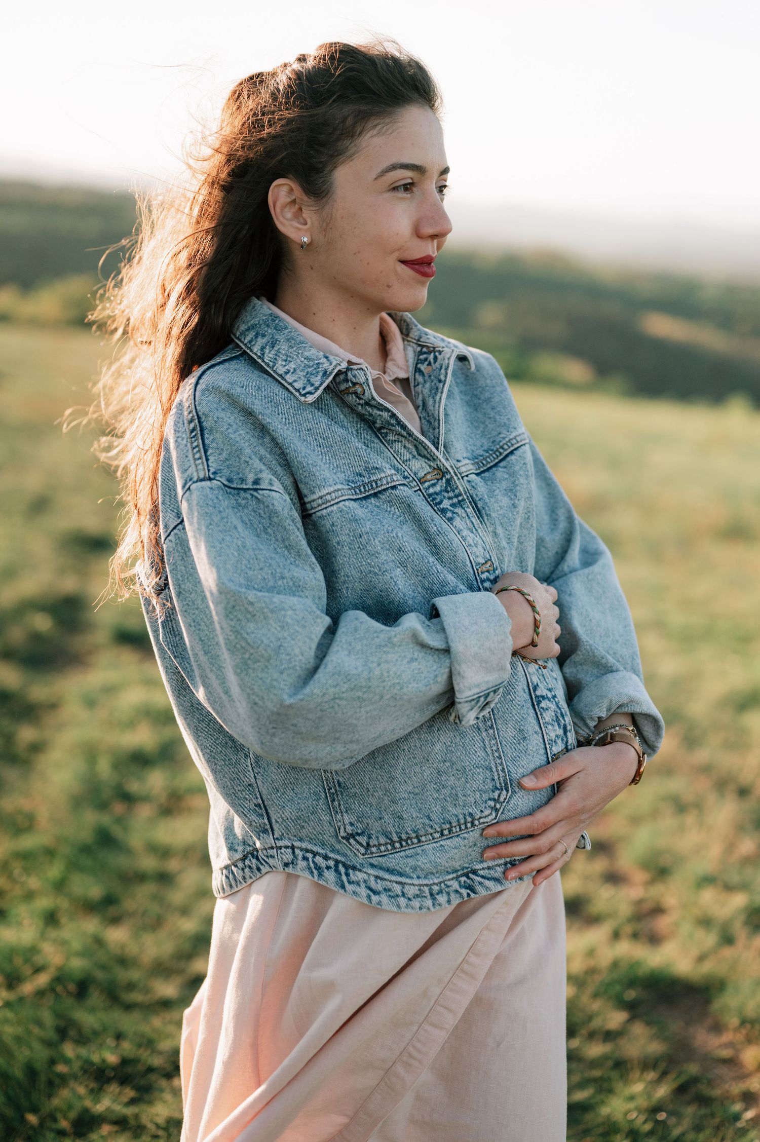 A person in a denim jacket and pink dress stands in a sunlit field during golden hour.