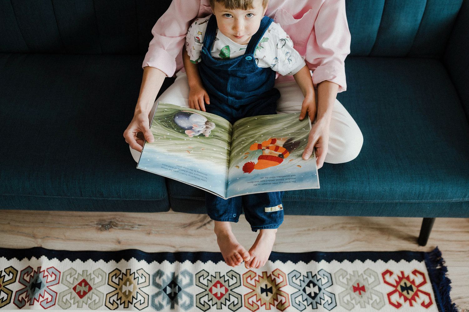 A child in denim overalls sits on a teal couch reading a picture book over a patterned rug.
