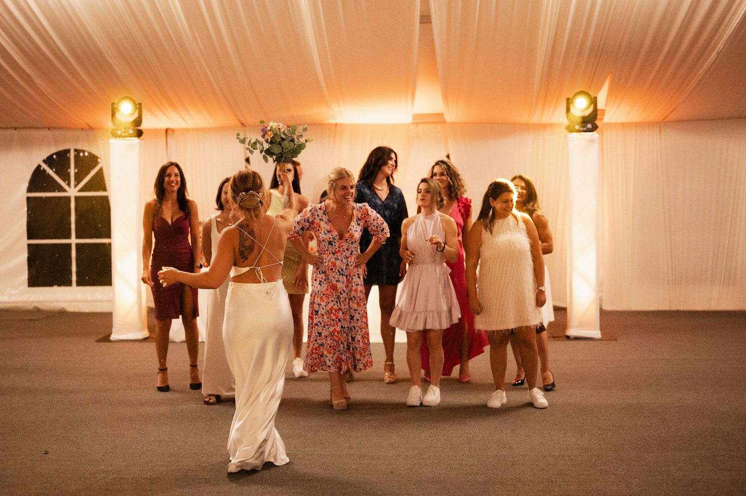 Wedding guests dance and celebrate during a reception in a warmly lit venue with white walls and wooden floors.