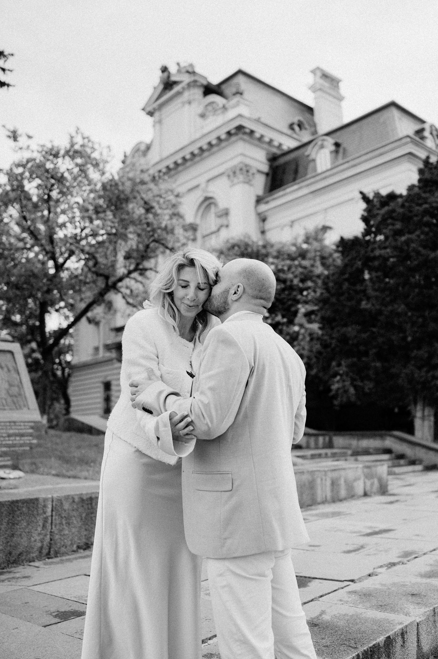 A romantic couple dressed in white embraces on stone steps with an elegant building in the background.