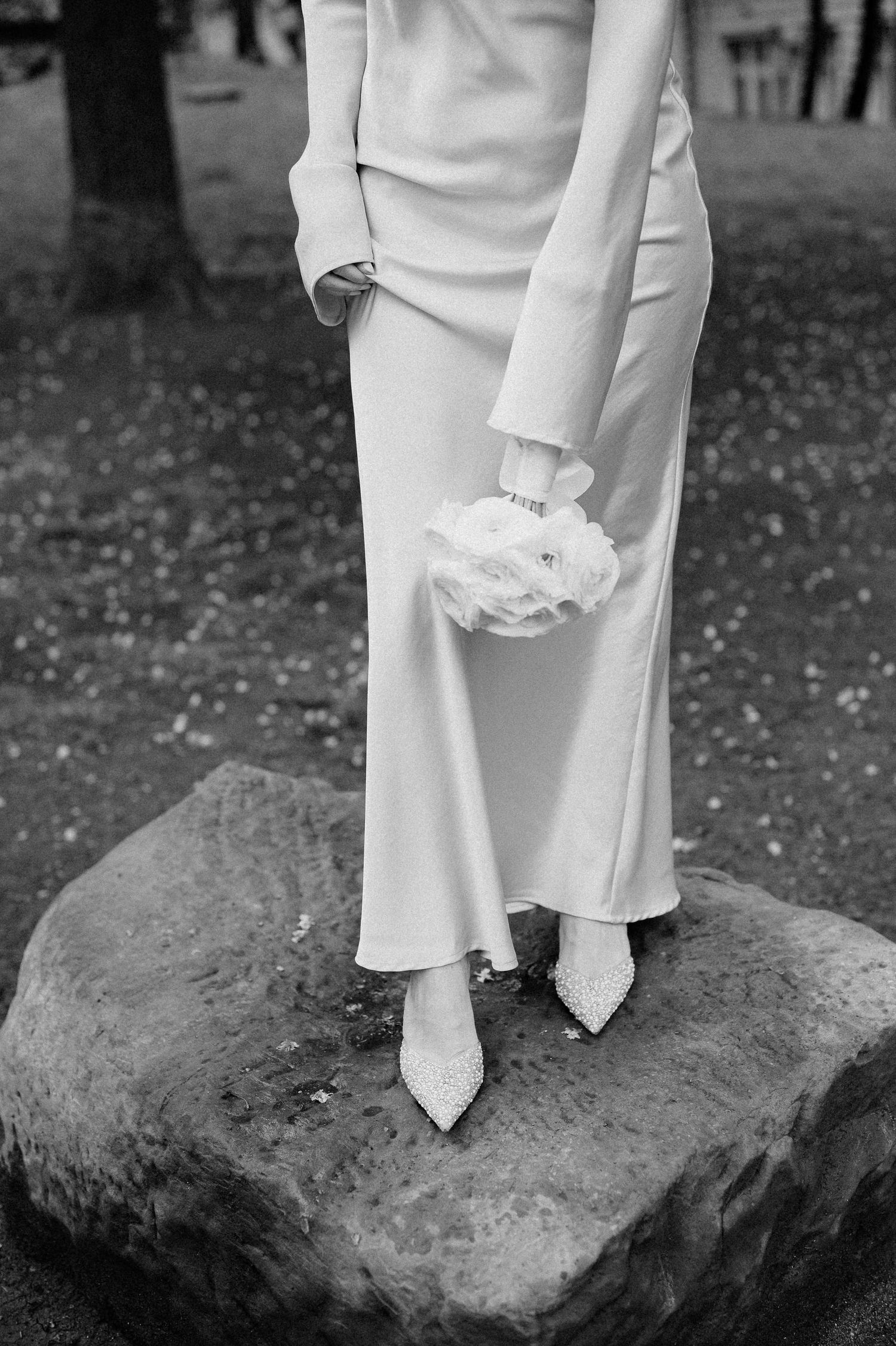 Black and white detail of formal white shoes and dress hem on a rocky surface.