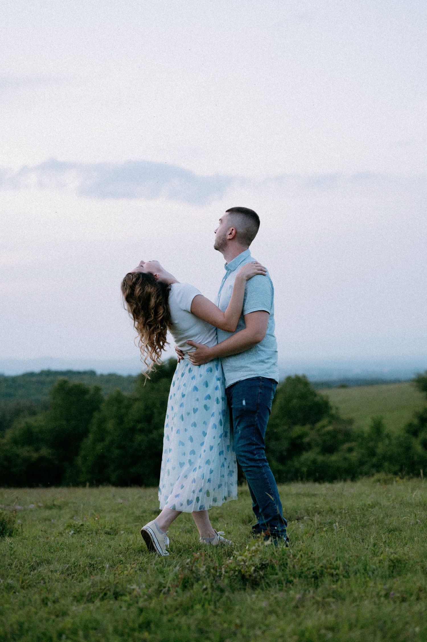 Two people share a romantic moment on a hilltop with mountains in the background at dusk.