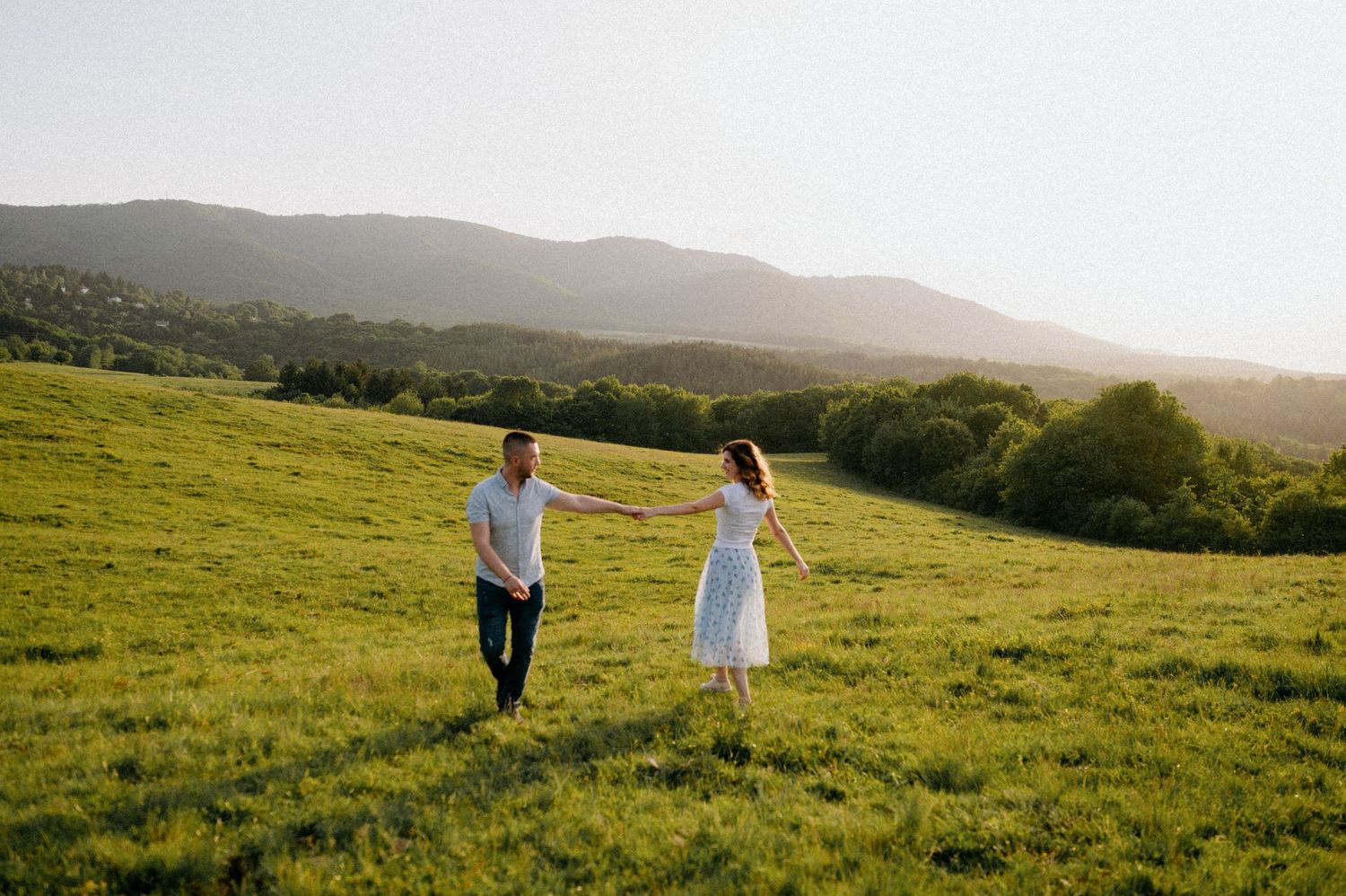 Two people walk hand in hand through a scenic hilltop meadow at golden hour.
