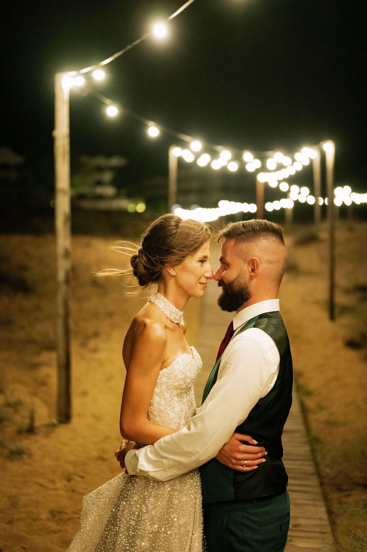 A romantic wedding couple embraces under string lights on a beach boardwalk at night.