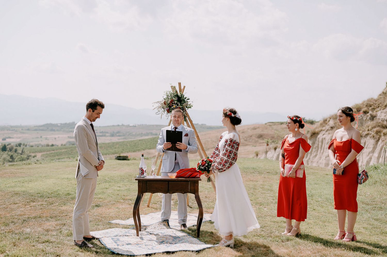 Wedding party in coral dresses and light suits gather on a grassy hillside for outdoor celebration.