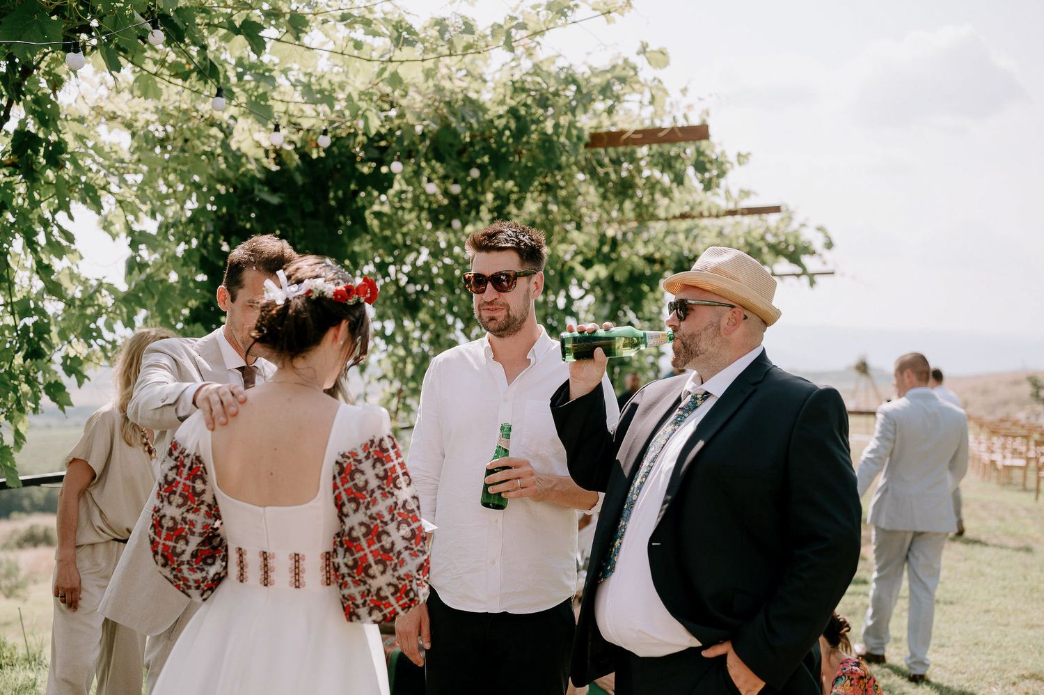 Outdoor wedding celebration with guests enjoying drinks under tree canopy.