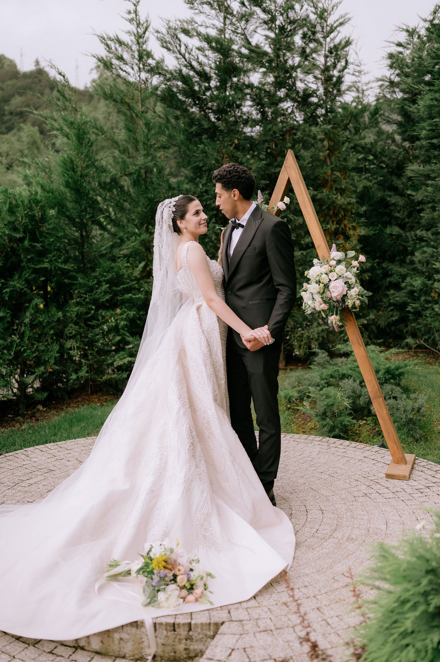 Elegant outdoor wedding ceremony setup with wooden arch and floral arrangements against evergreen trees.