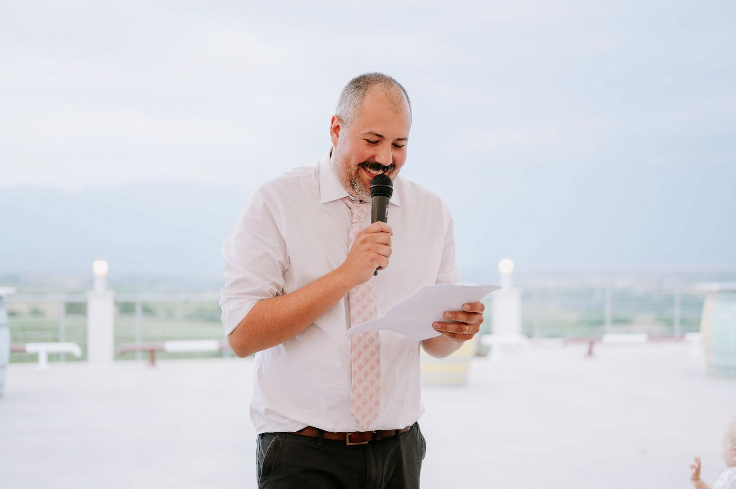 Speaker delivers remarks at an outdoor rooftop wedding venue against a bright sky.