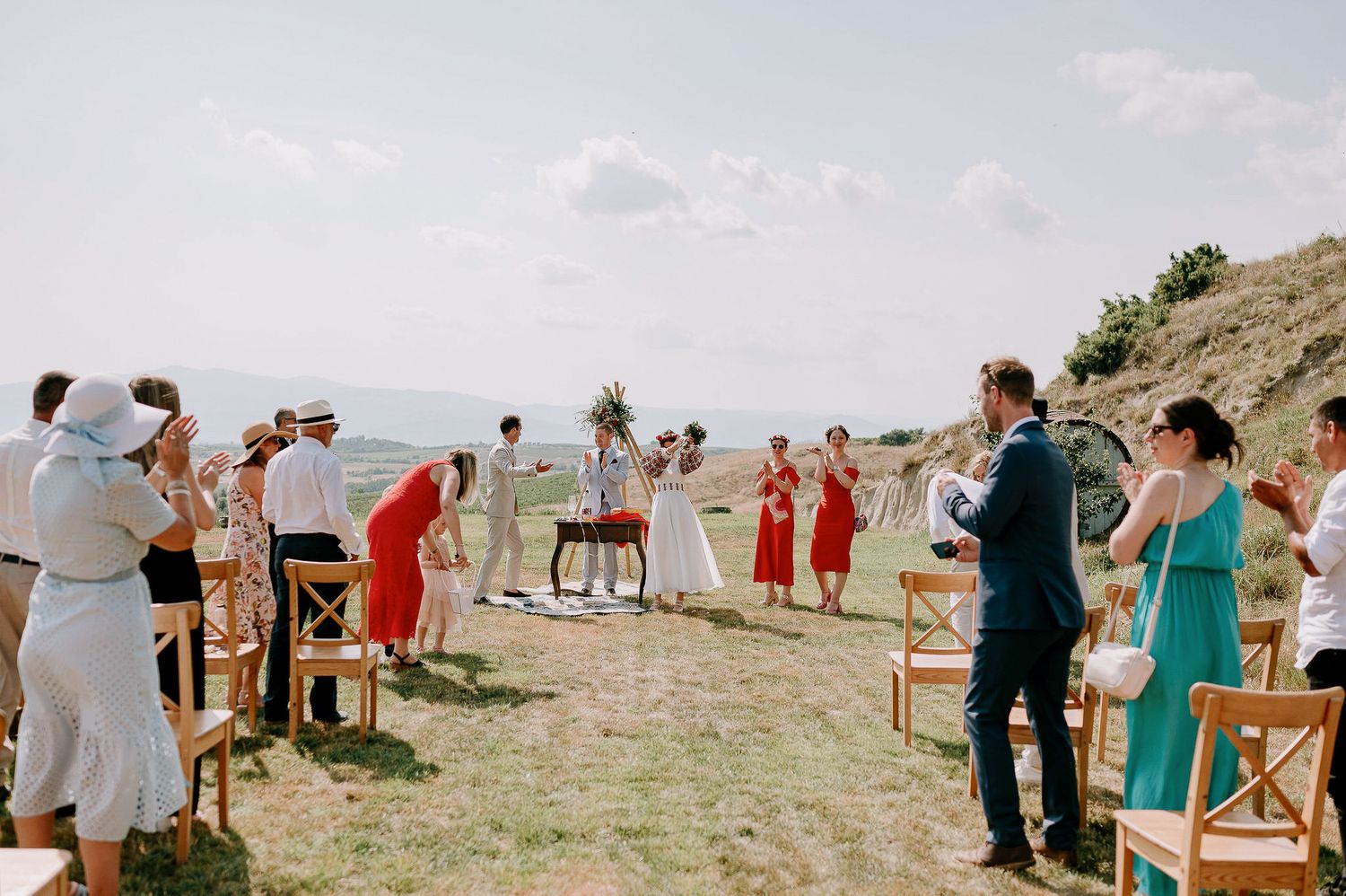 Wedding guests gather on a grassy hillside with wooden chairs set up for an outdoor ceremony.