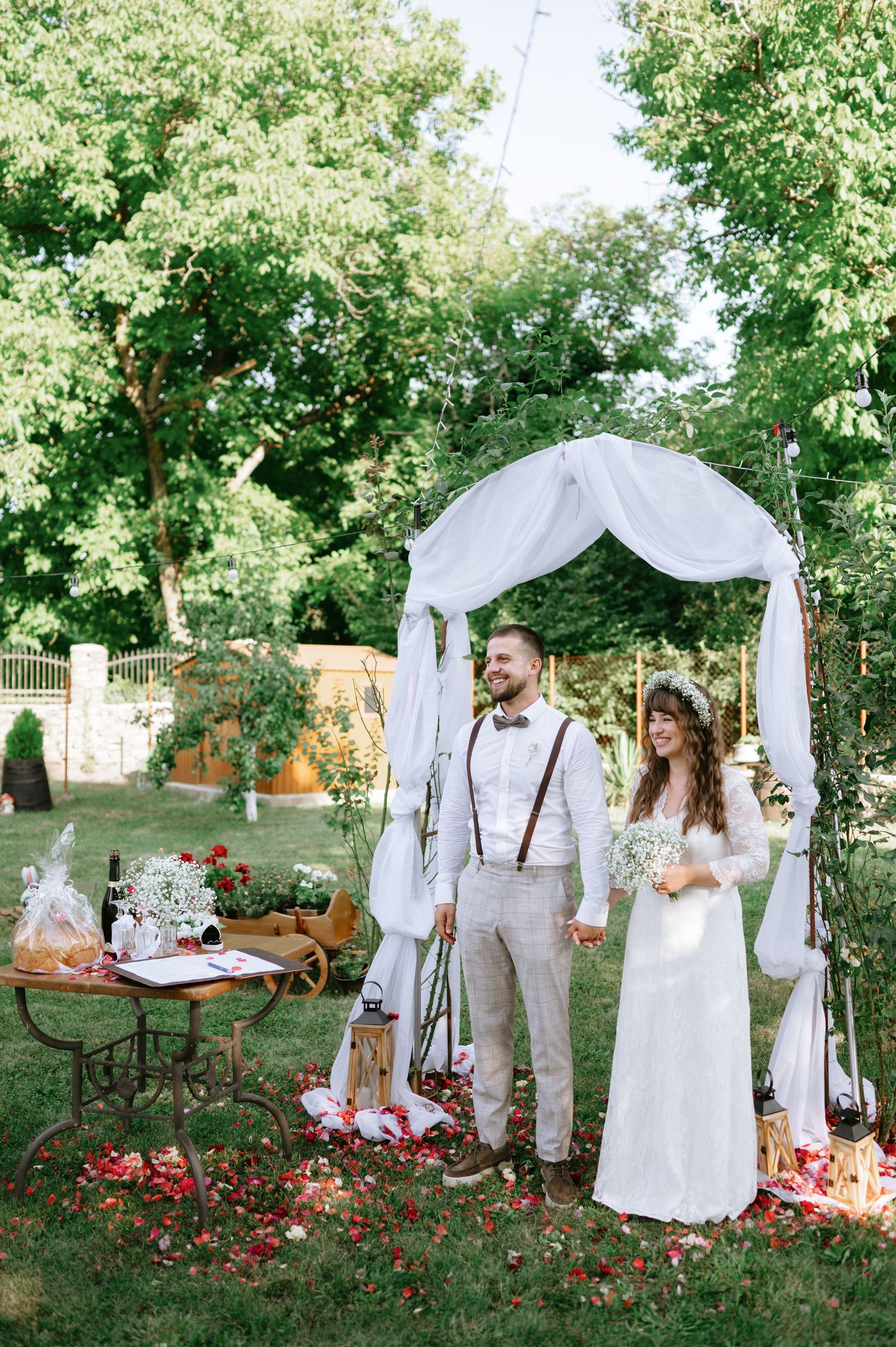 Intimate garden wedding setup with white draped arch and vintage furniture under green canopy.