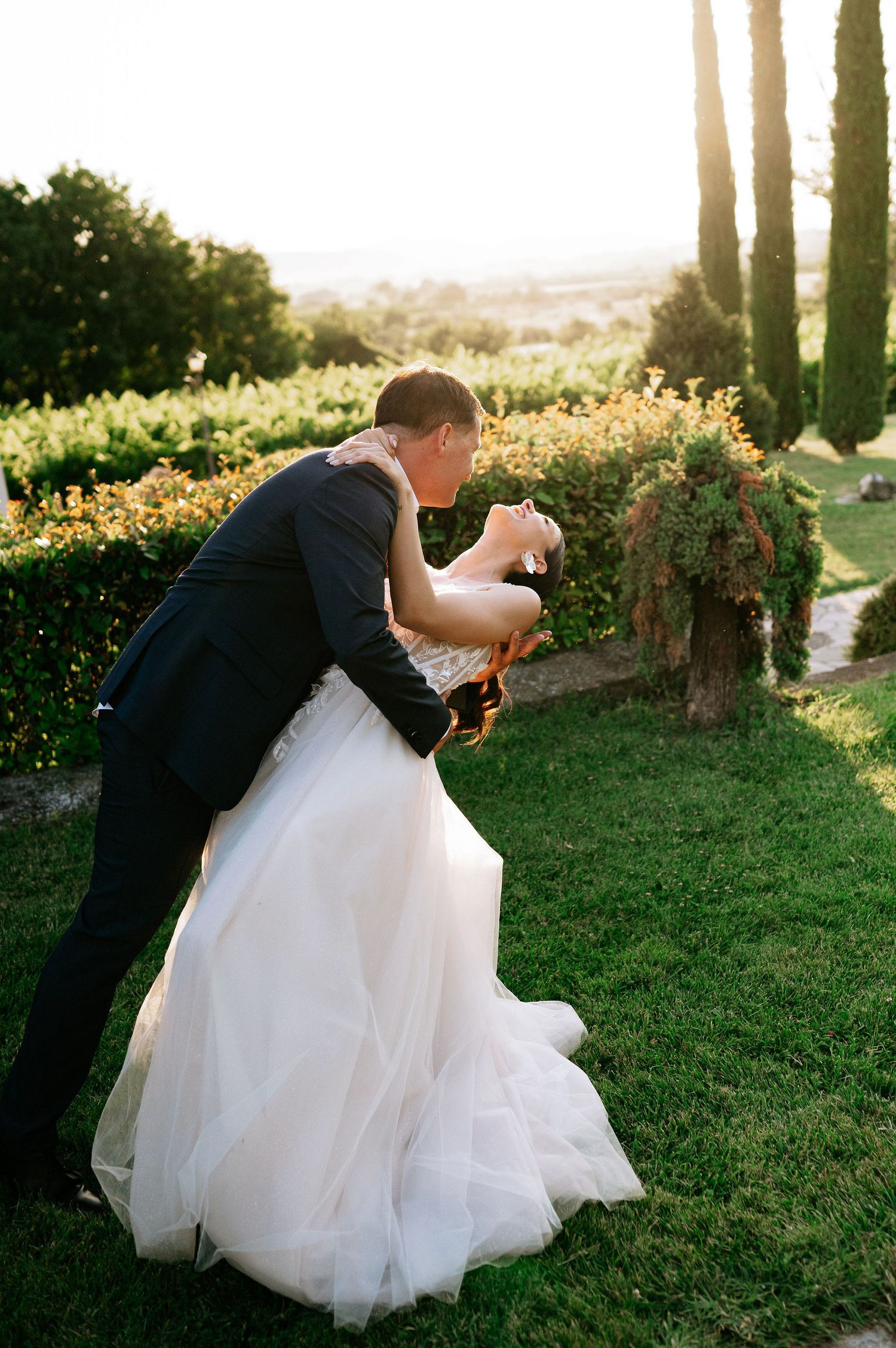 Dramatic dip pose between newlyweds in garden setting with cypress trees.