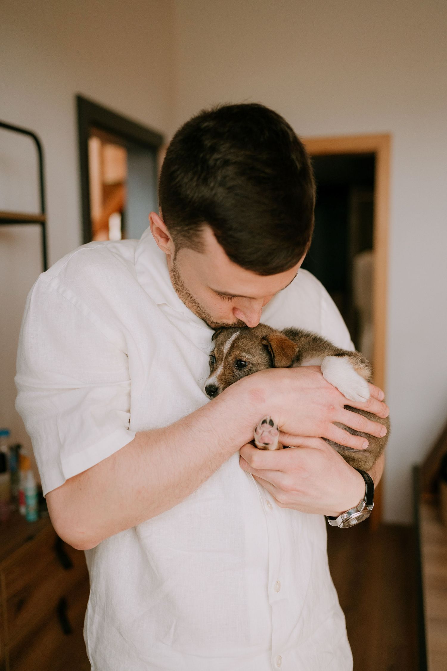 A couple in white clothing shares a tender moment with their small brown dog in a rustic wooden room.