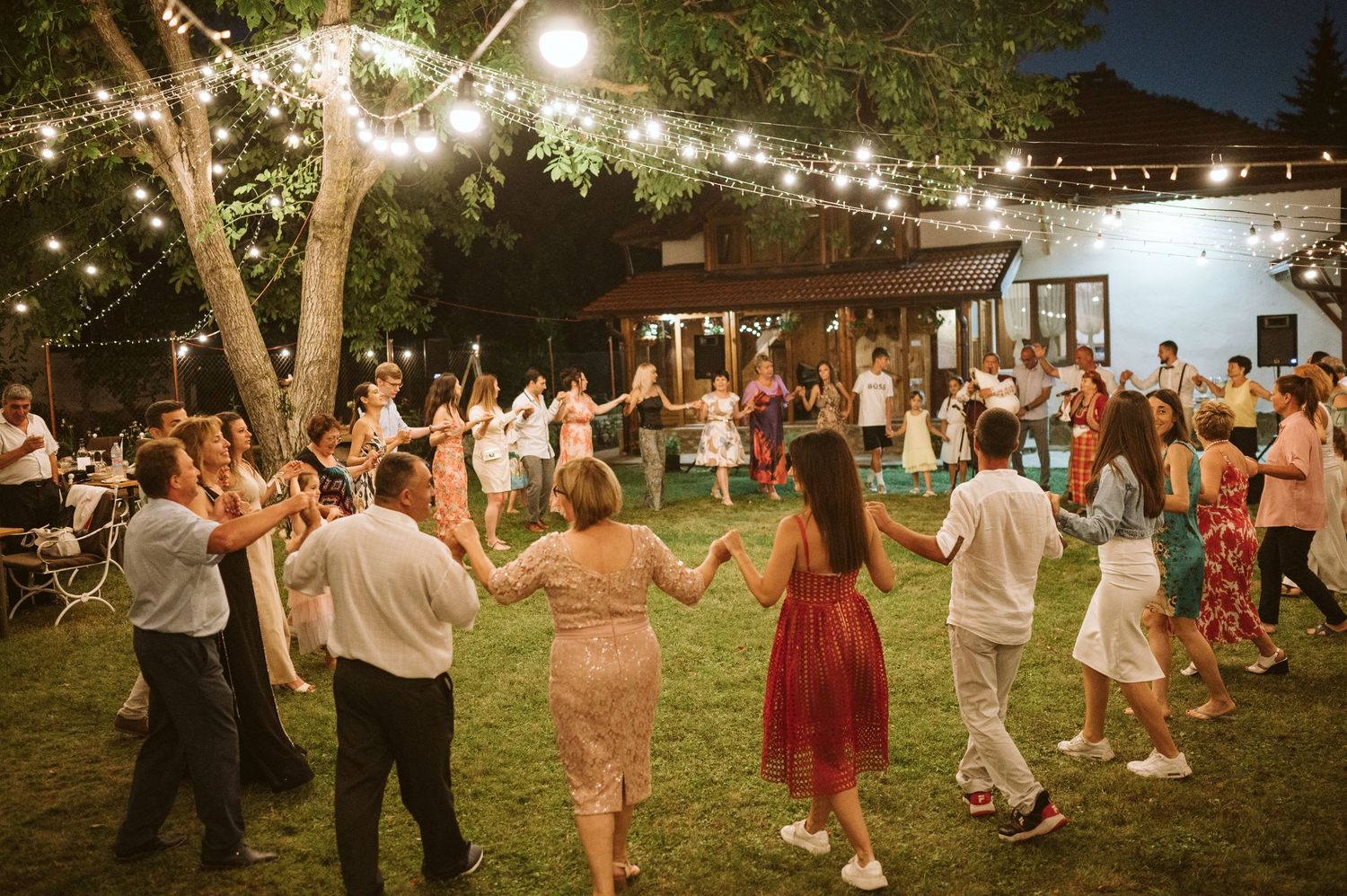Guests dance in a line formation on grass under strung lights at outdoor wedding reception.