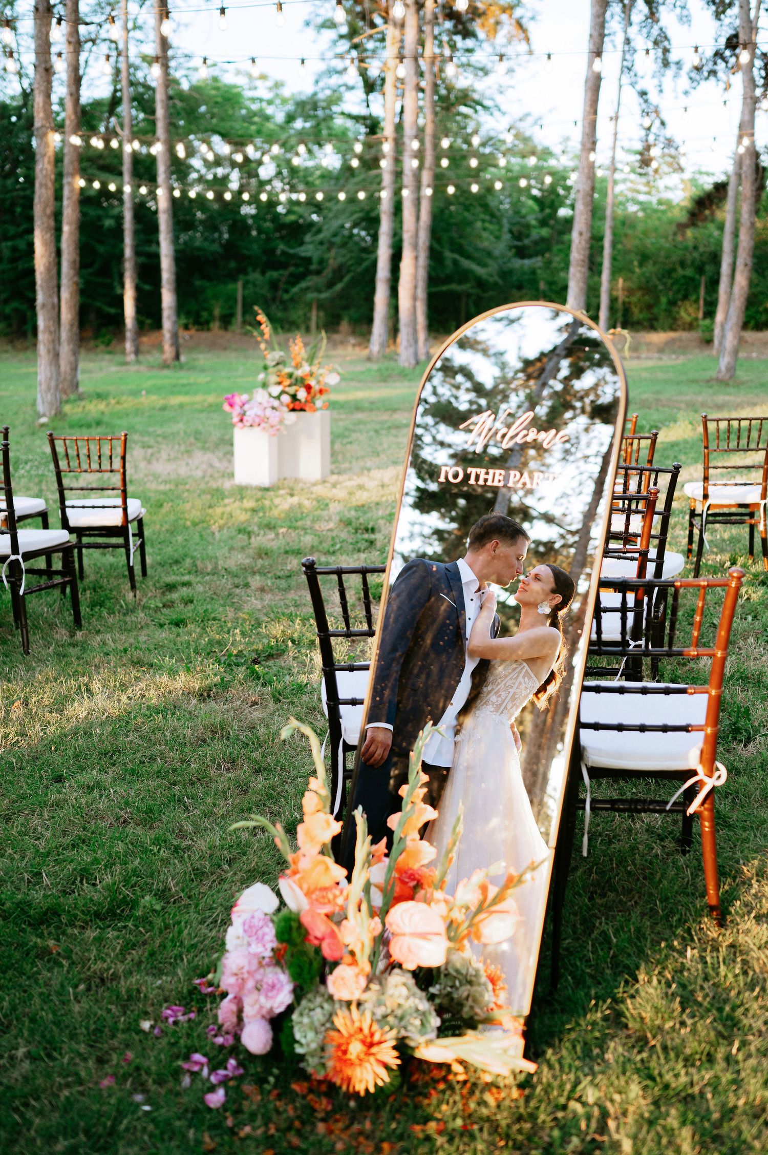 Rustic woodland wedding ceremony site decorated with string lights and colorful floral arrangements.