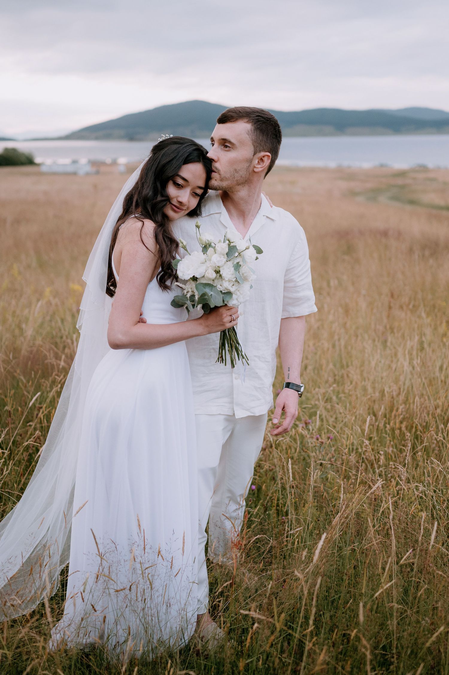 A couple in white wedding attire poses lovingly in tall grass with a scenic mountain backdrop.