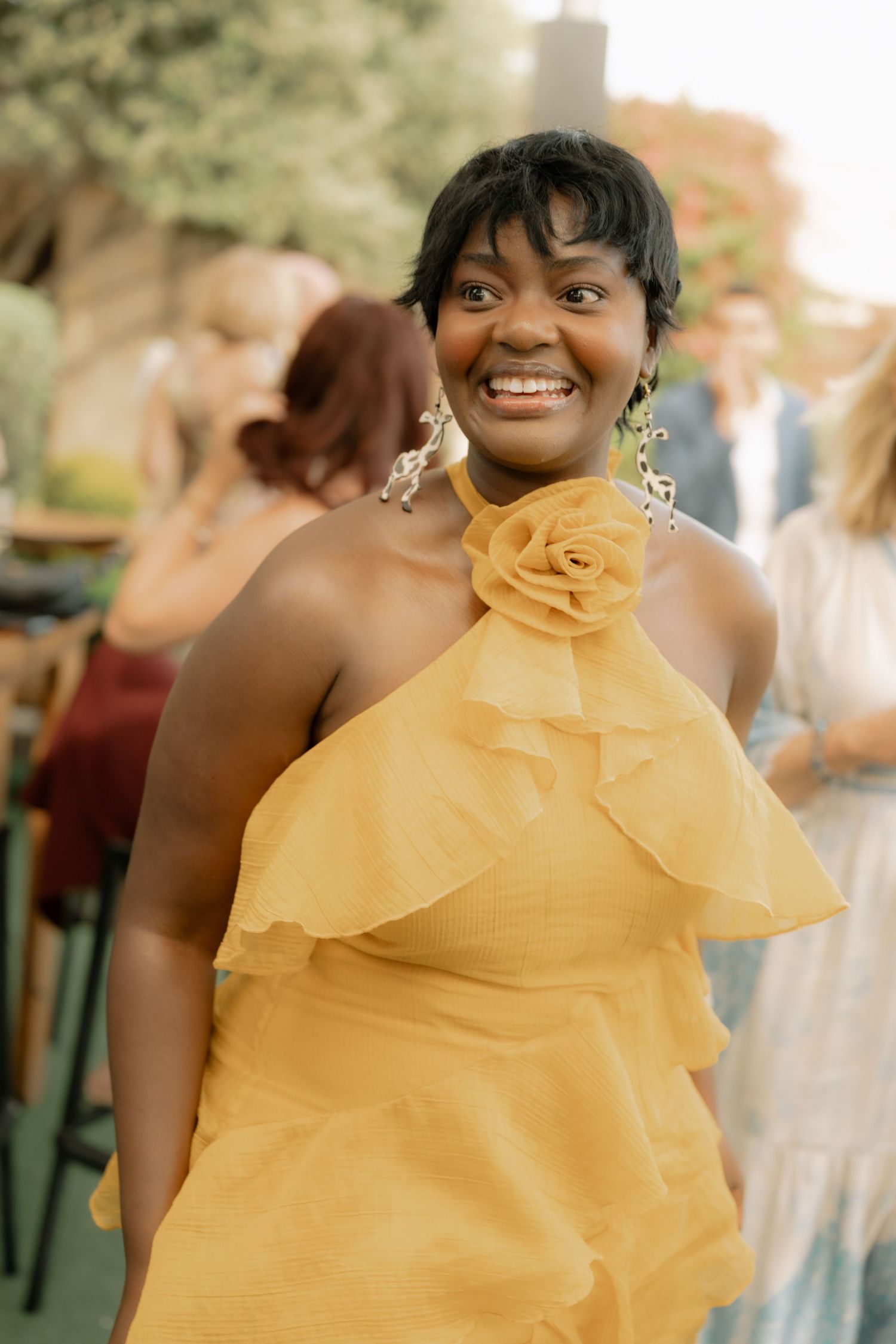 Smiling woman in yellow ruffled halter dress with floral detail at outdoor event.