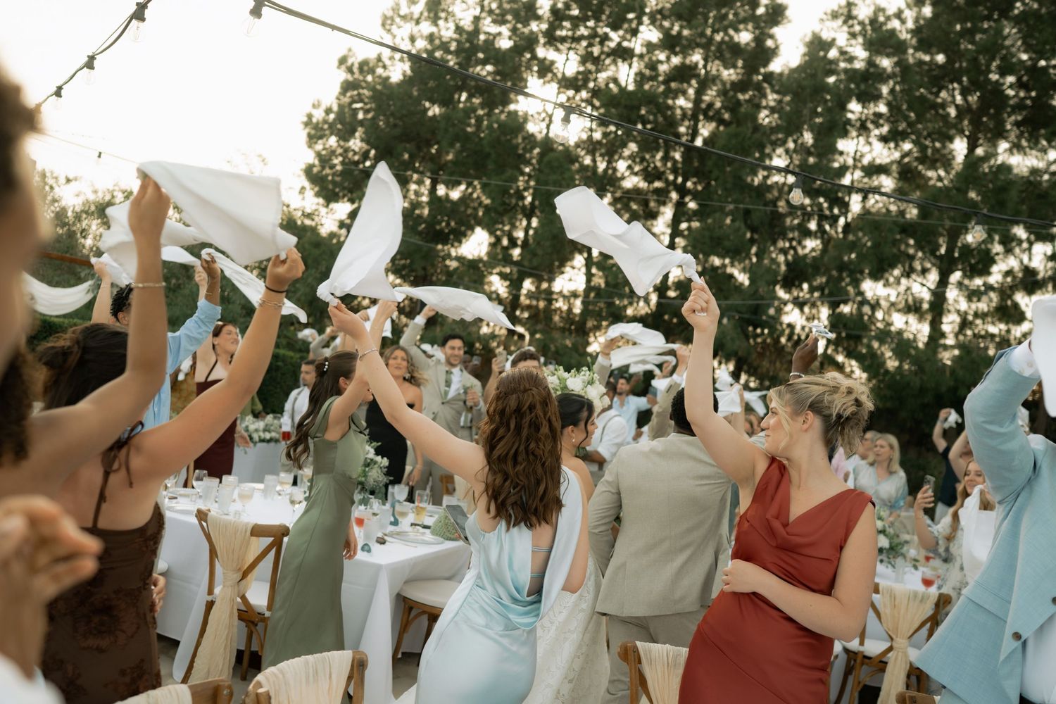 Wedding guests waving white napkins at an outdoor reception dinner party surrounded by trees and decorated tables.