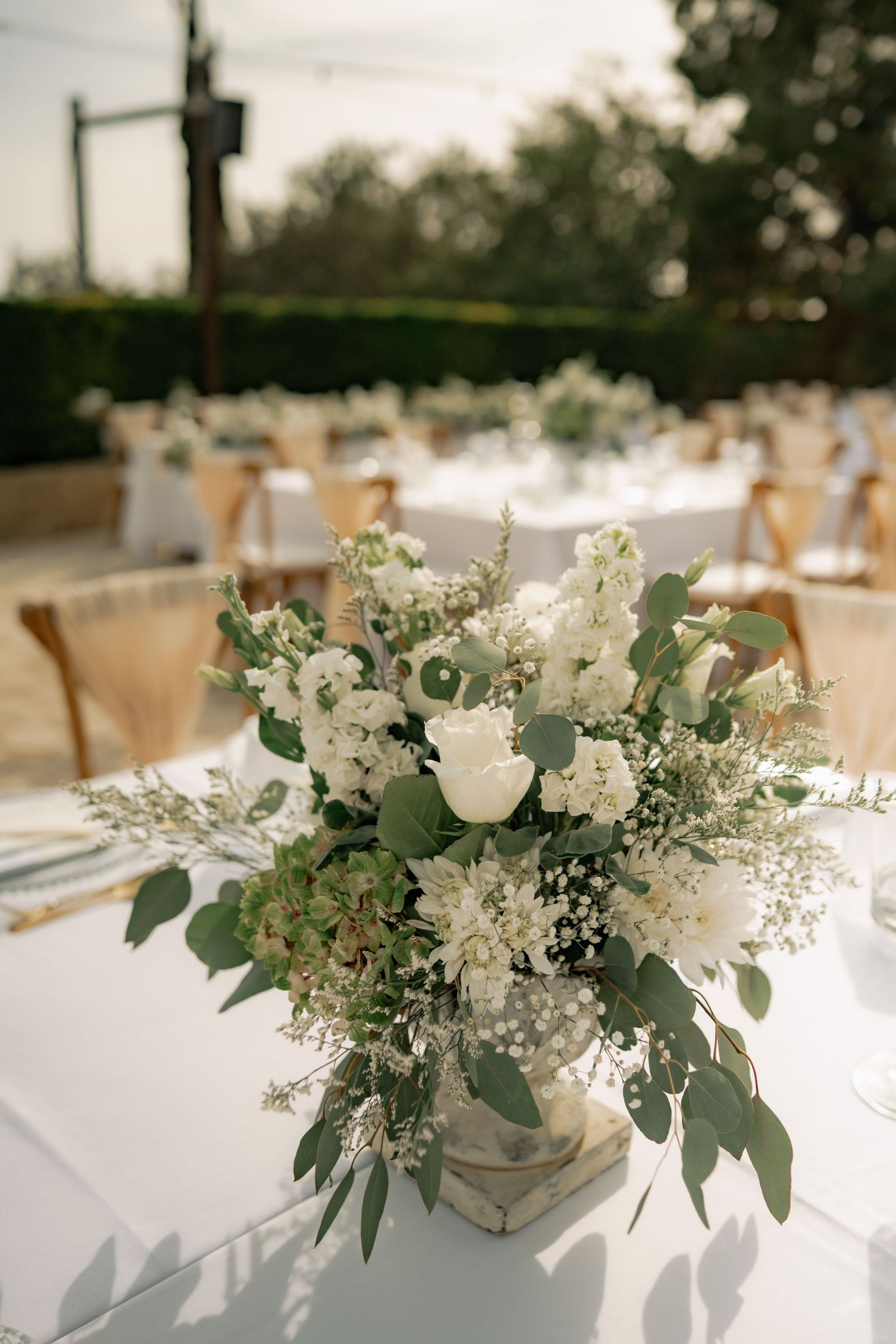 White floral centerpiece with eucalyptus greenery on white tablecloth at an outdoor wedding reception.