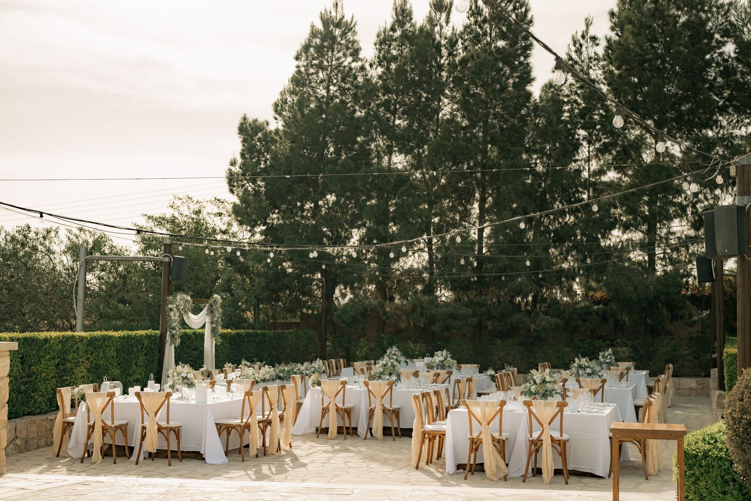 Elegant outdoor wedding reception with white tablecloths, wooden chairs, greenery centerpieces, and string lights among tall trees.