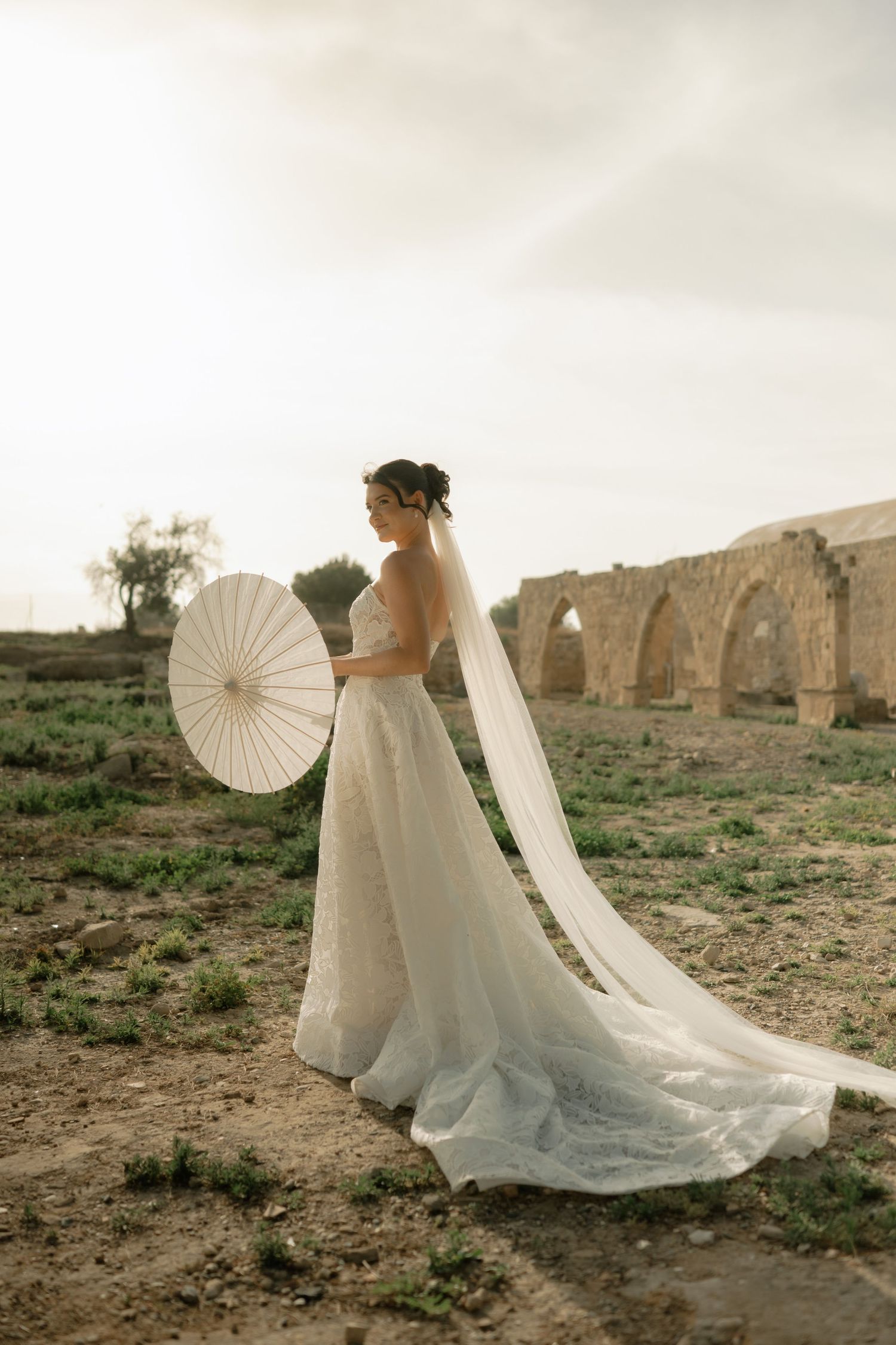 Bride in white lace gown with long veil holds parasol near ancient stone arches outdoors.